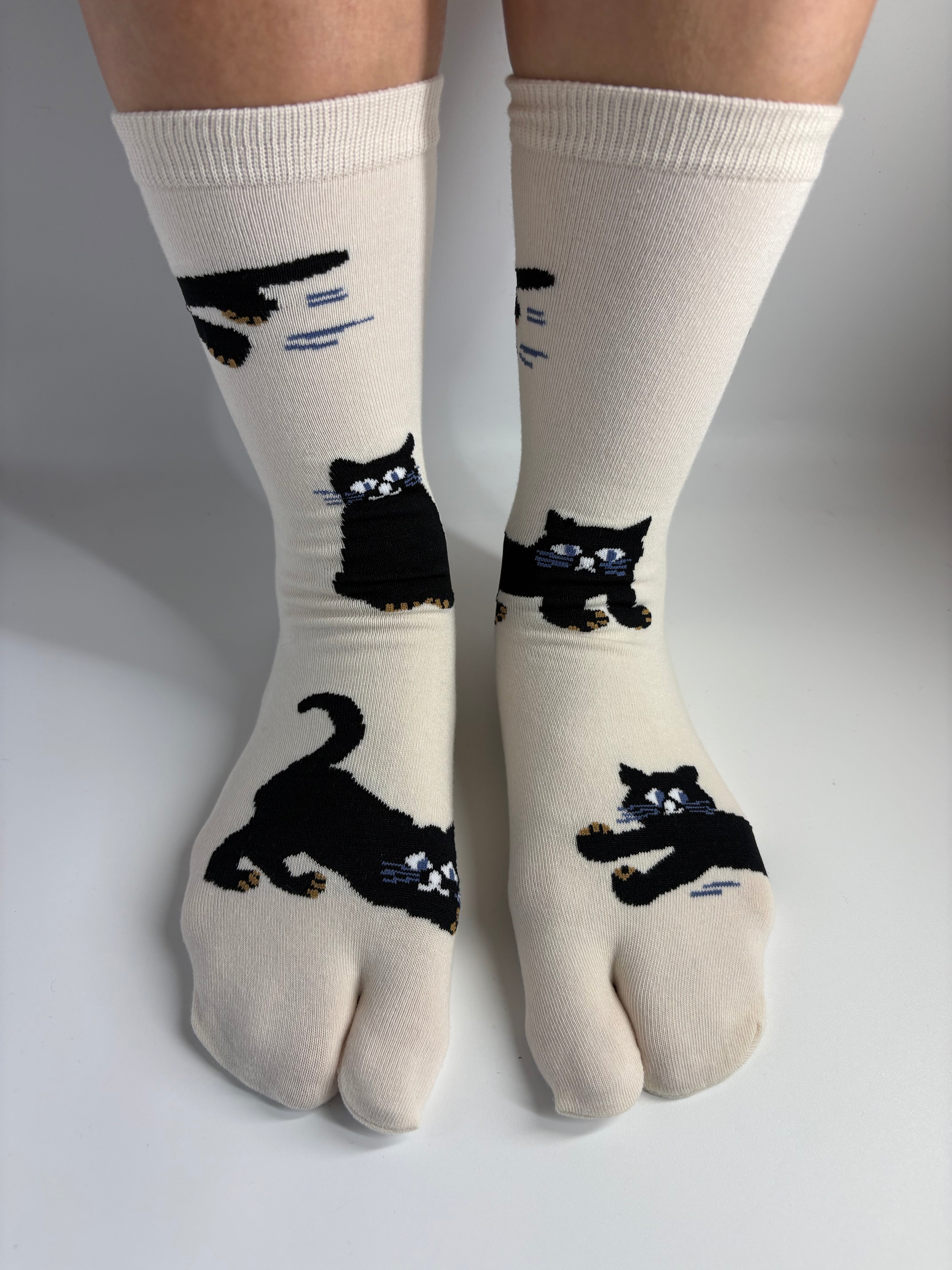 A pair of off-white crew socks with black cat illustrations, worn on feet against a plain light gray background.
