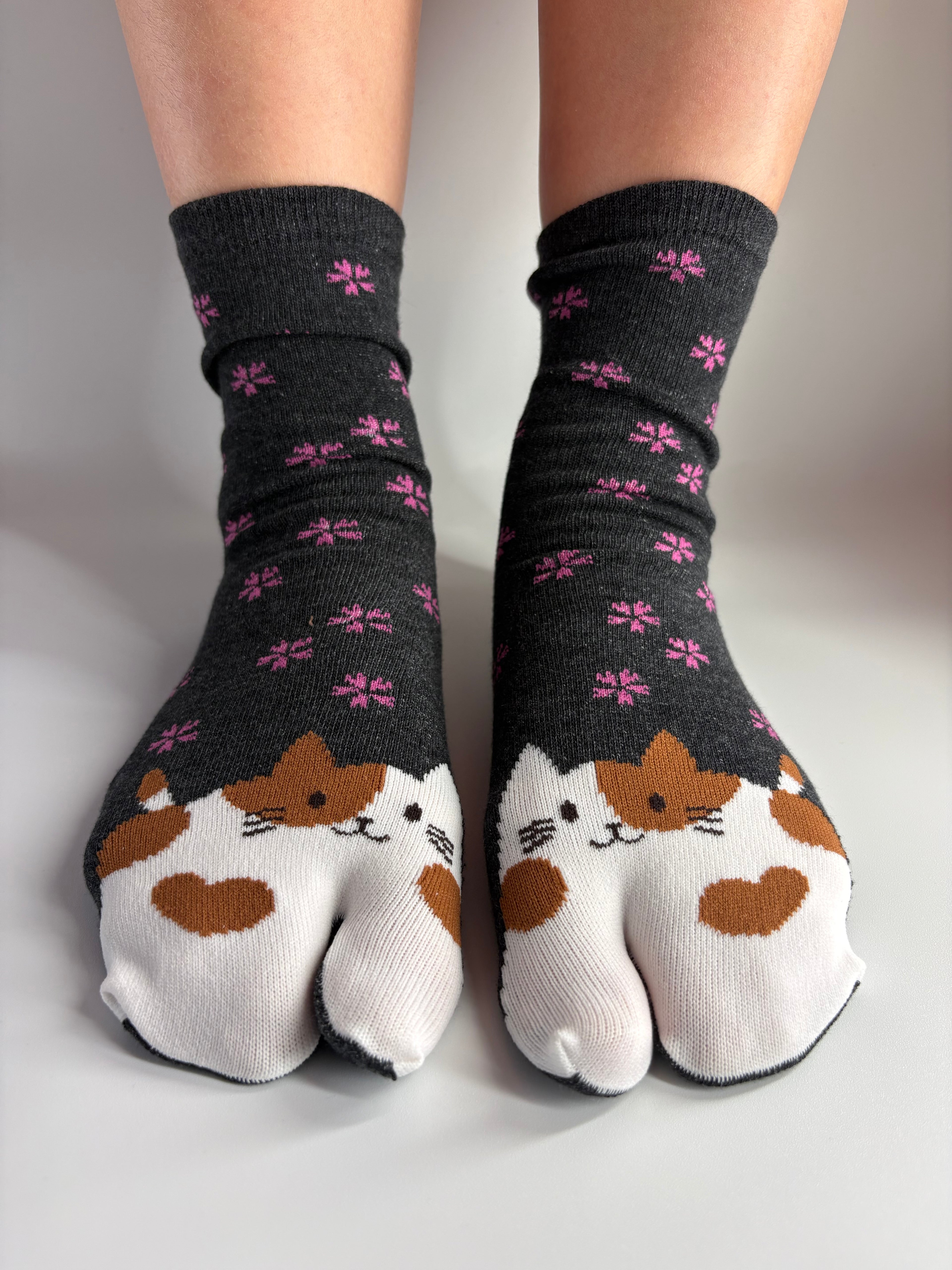 A pair of split-toe socks worn on feet, featuring a white and brown calico cat face design on the toes and pink flower patterns on a dark charcoal gray background.
