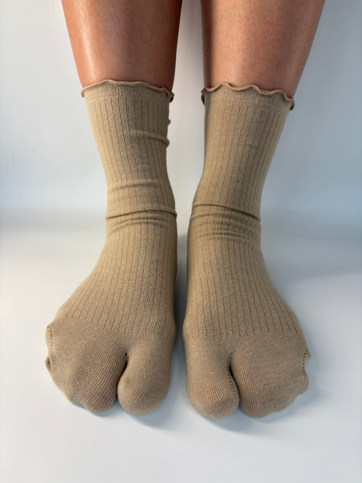A pair of beige ribbed tabi socks with ruffle edges worn on feet, shown against a plain light gray background.