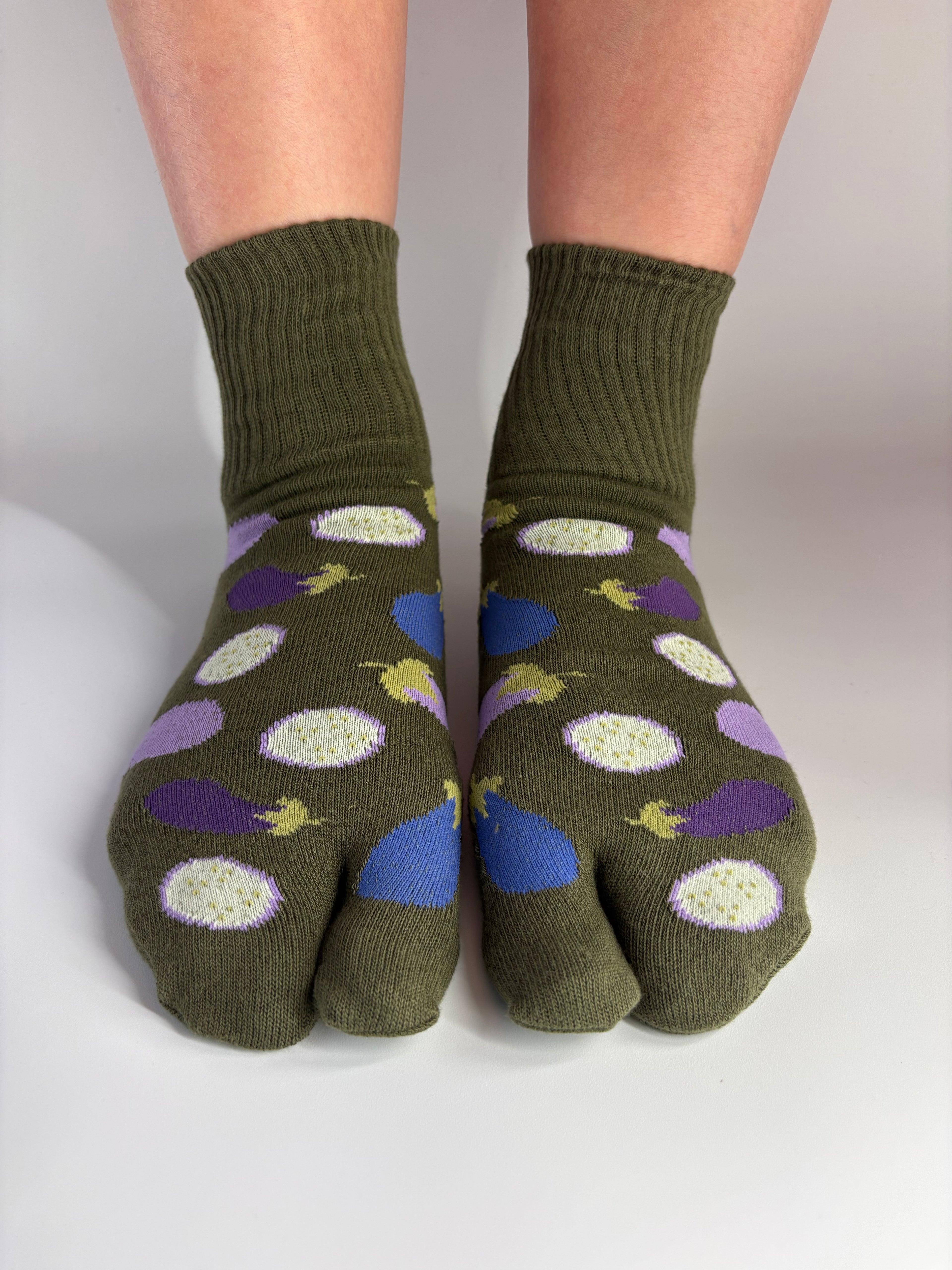 A pair of olive green tabi socks with a colorful eggplant pattern, worn on feet against a neutral light gray background.