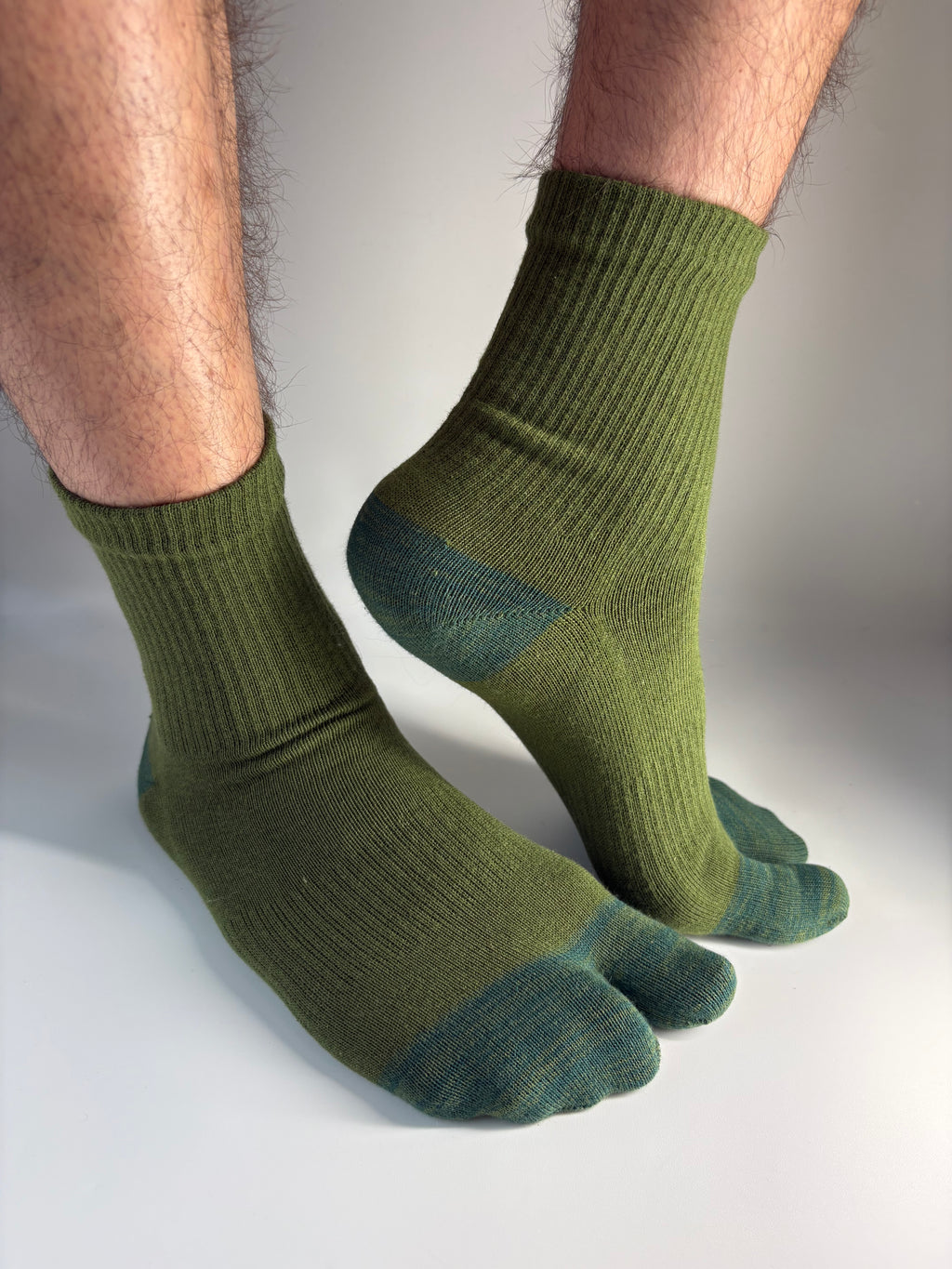 A pair of olive green tabi split-toe socks worn on a person's feet, shown against a neutral light gray background.