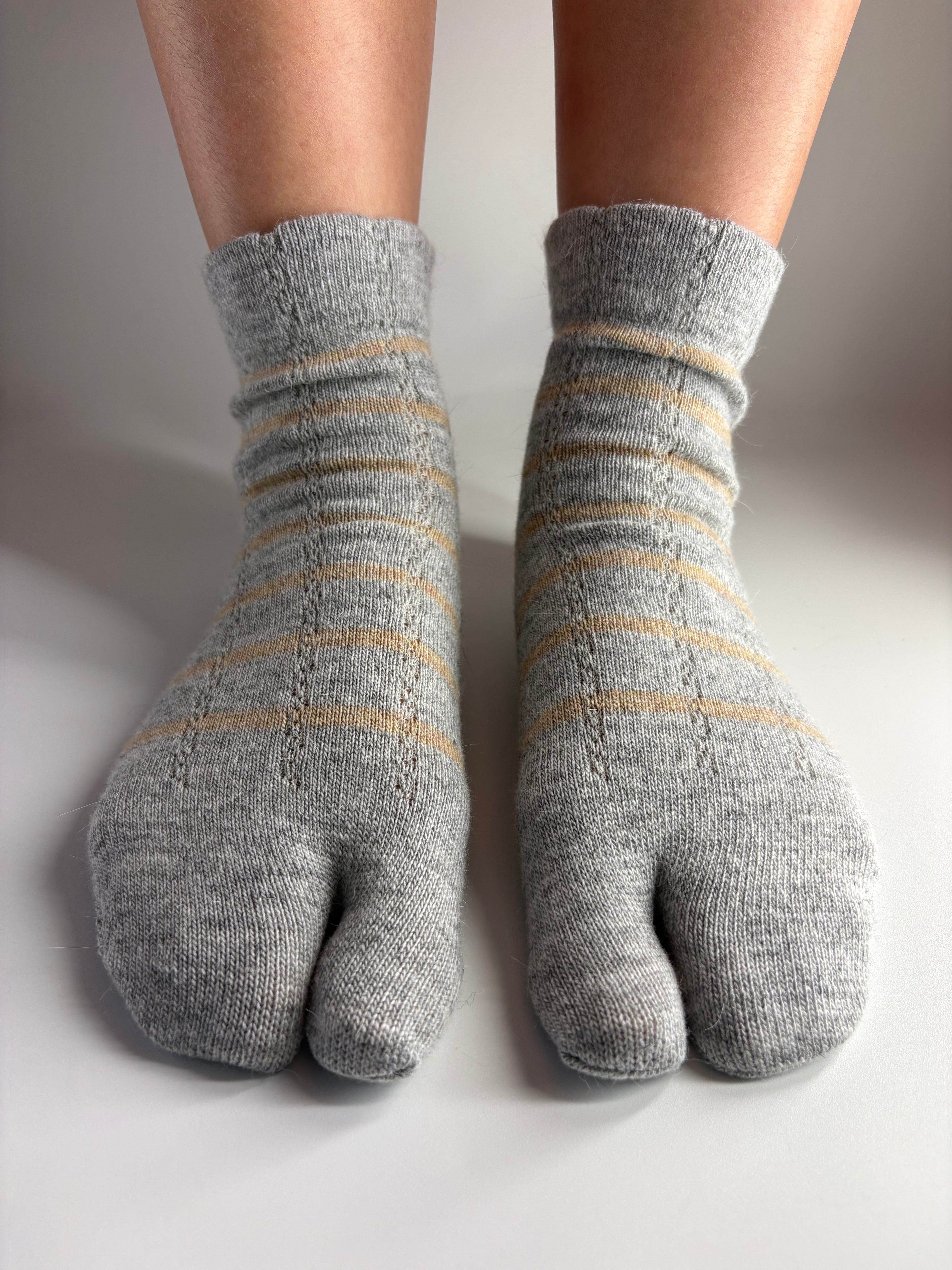 A pair of gray tabi socks with thin beige stripes, shown worn on a person's feet against a plain light gray background.