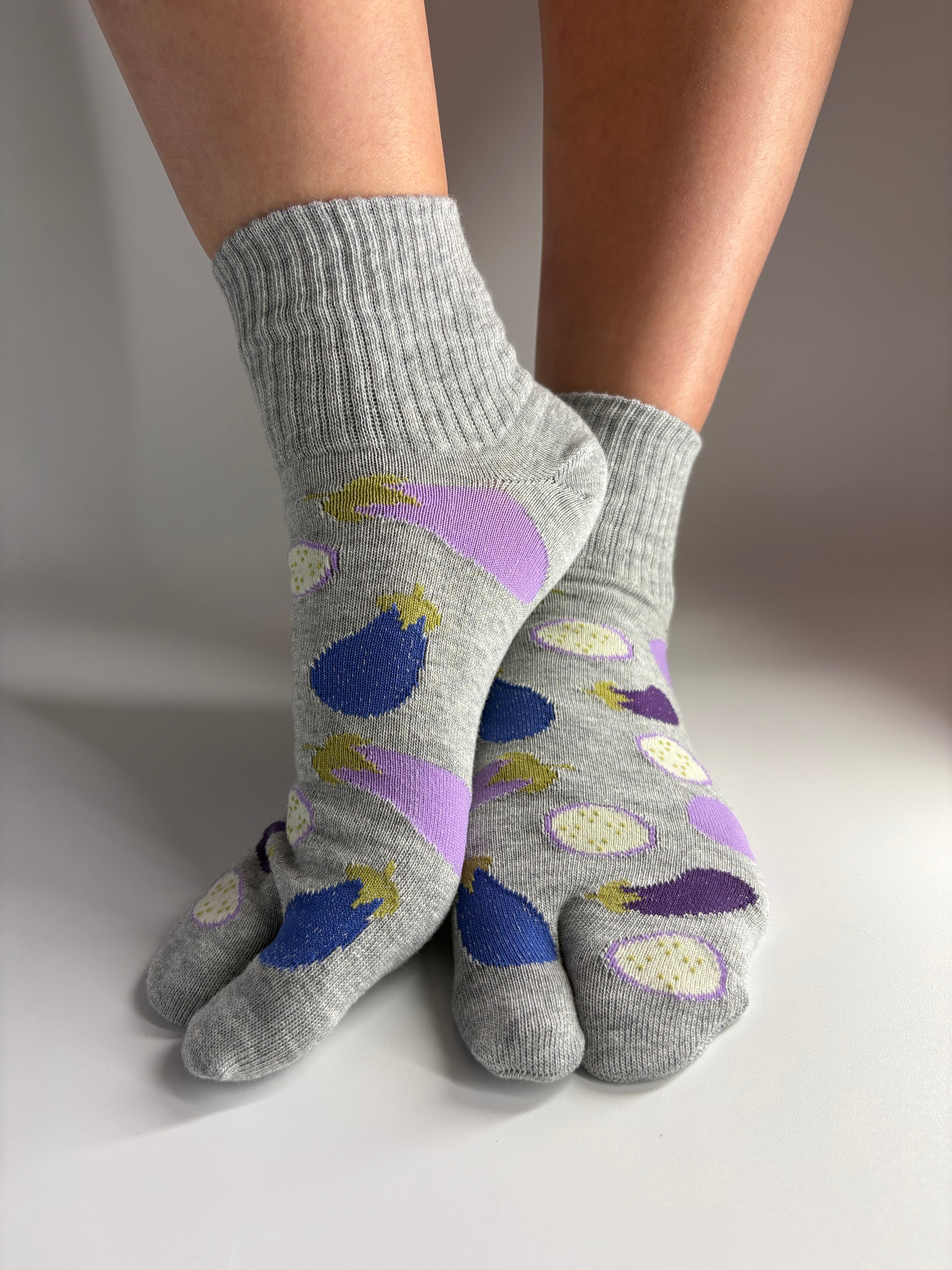 A pair of gray ribbed crew socks with a colorful eggplant pattern, worn on feet against a plain light gray background.