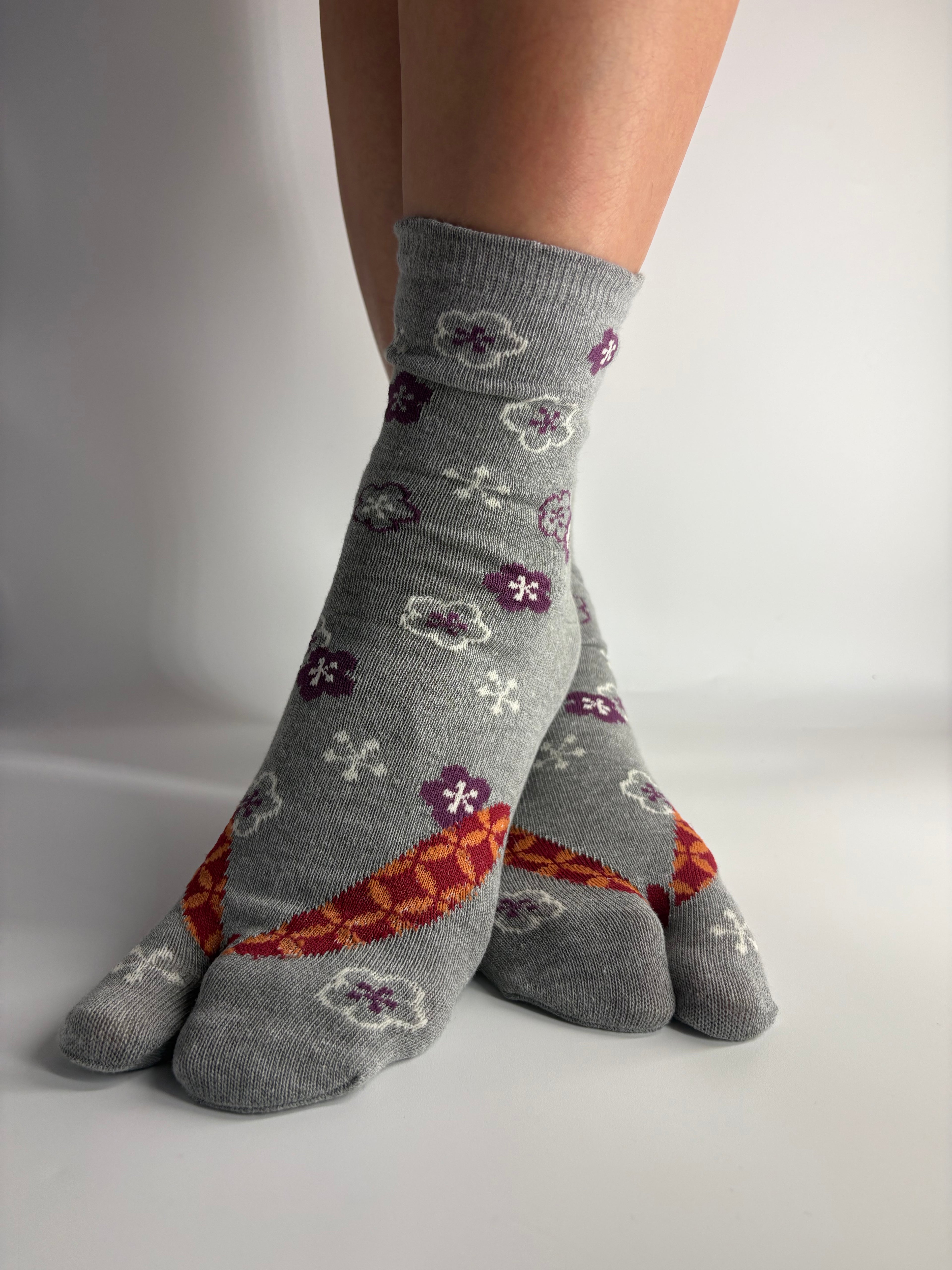 A pair of grey crew socks with a pattern of purple flowers, white snowflakes, and an orange geometric design, shown on a person's feet against a white background.