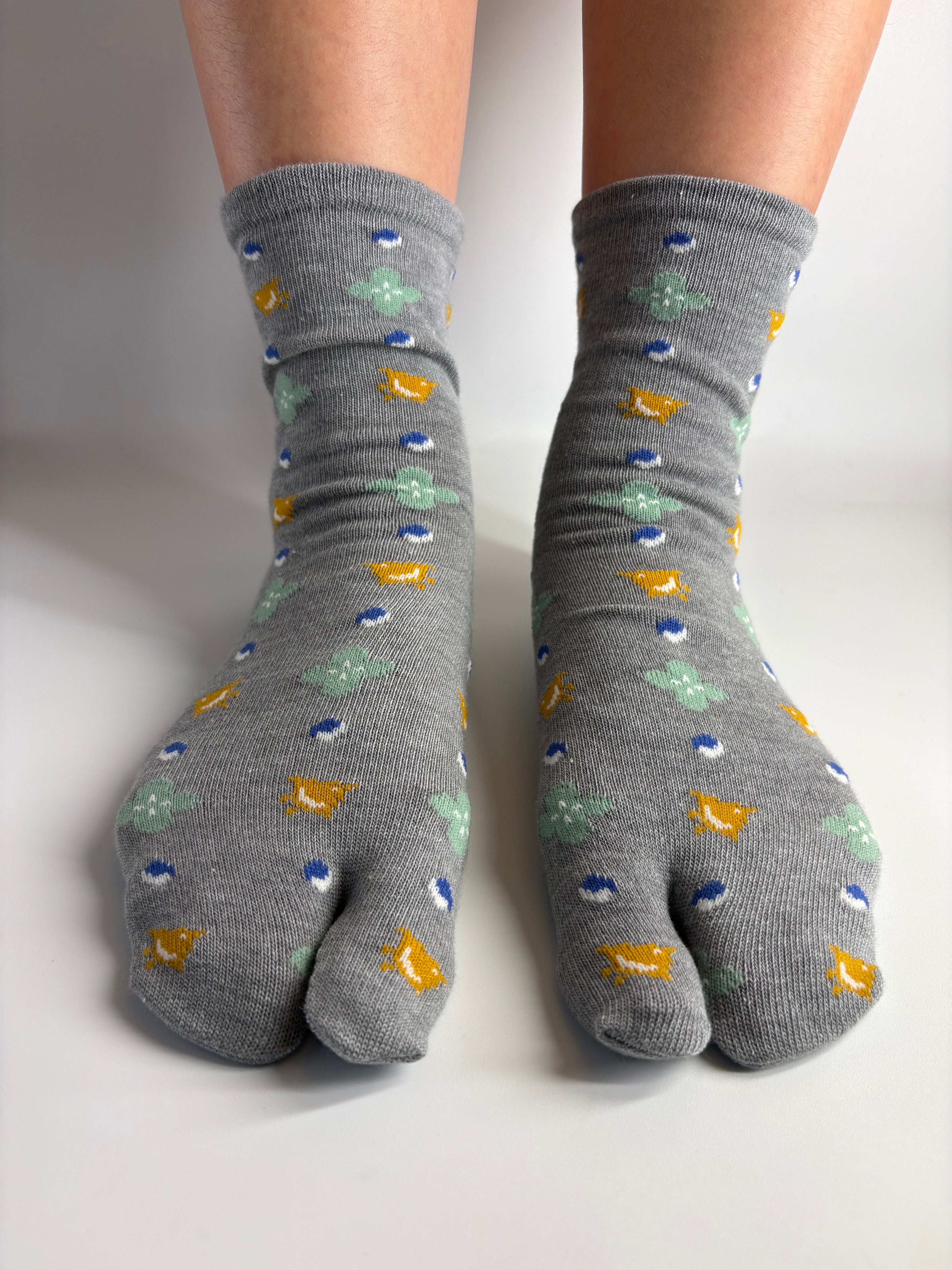 A pair of grey tabi socks with a colorful pattern of small yellow, blue, and mint green motifs, shown on a person's feet against a plain light background.