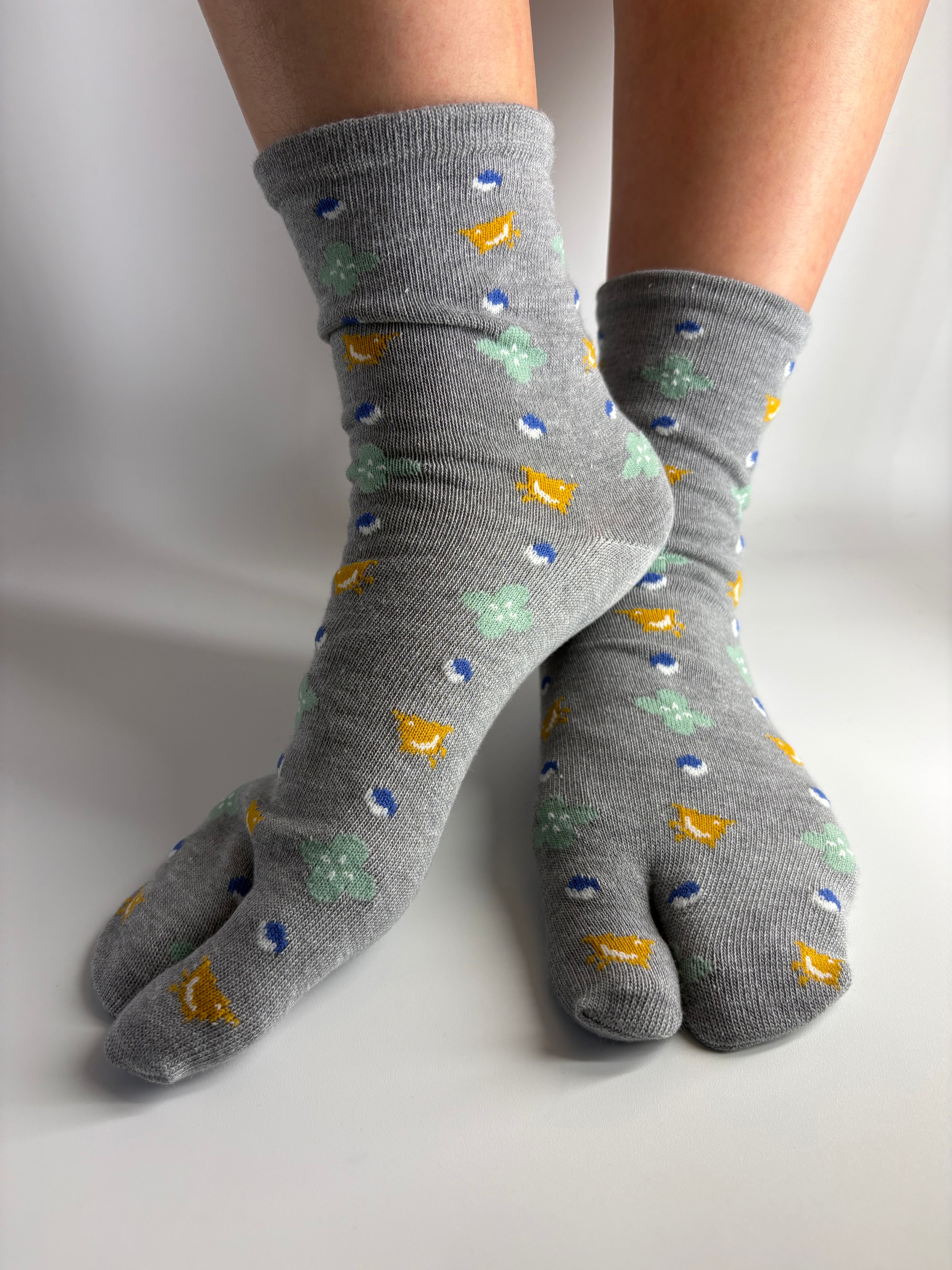 A pair of gray tabi socks with a colorful pattern of small yellow, blue, and mint green shapes, shown on a person's feet against a plain white background.