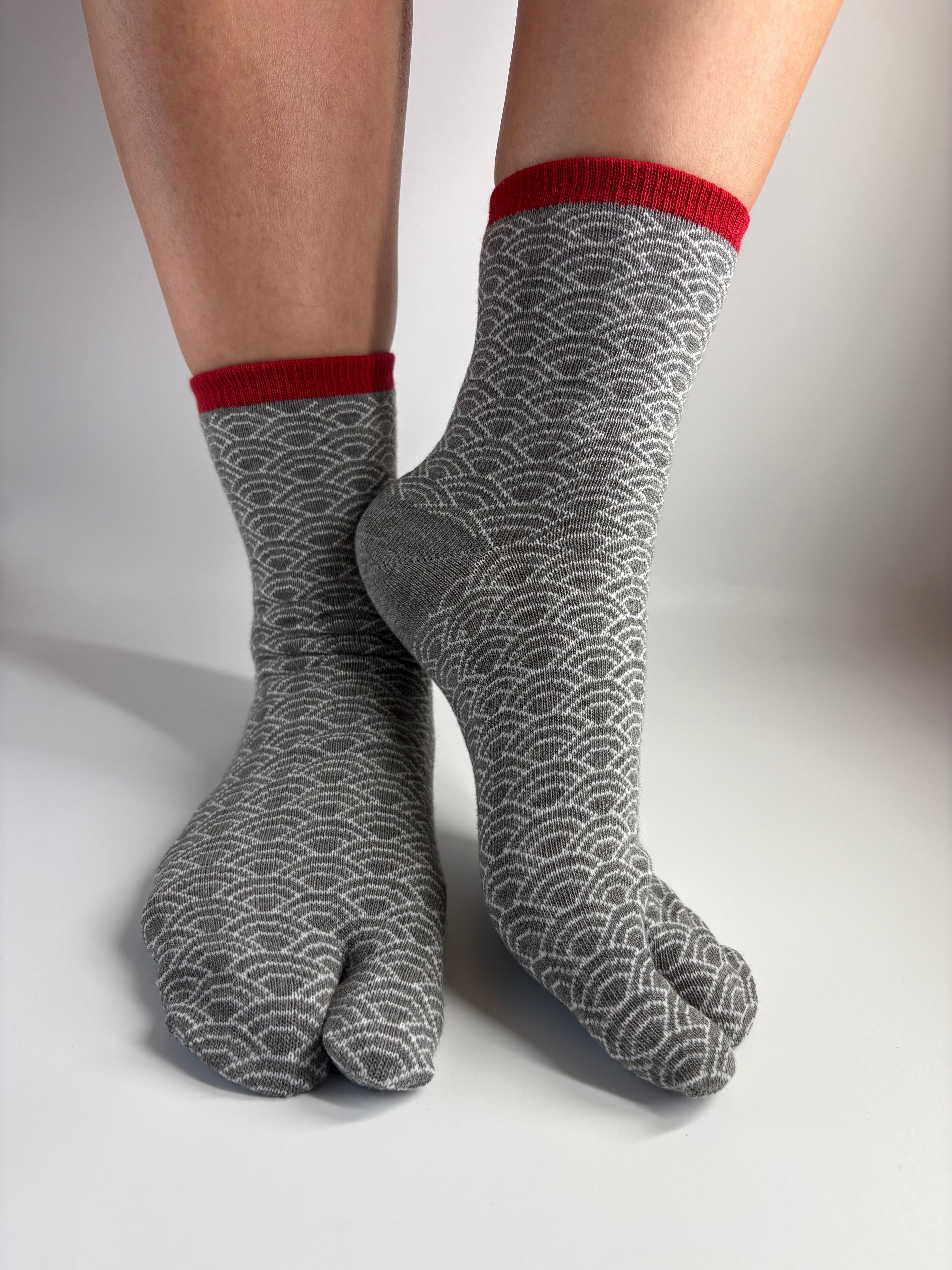 Gray tabi socks with a repeating wave pattern and red cuffs, shown on a person's feet against a neutral studio background.