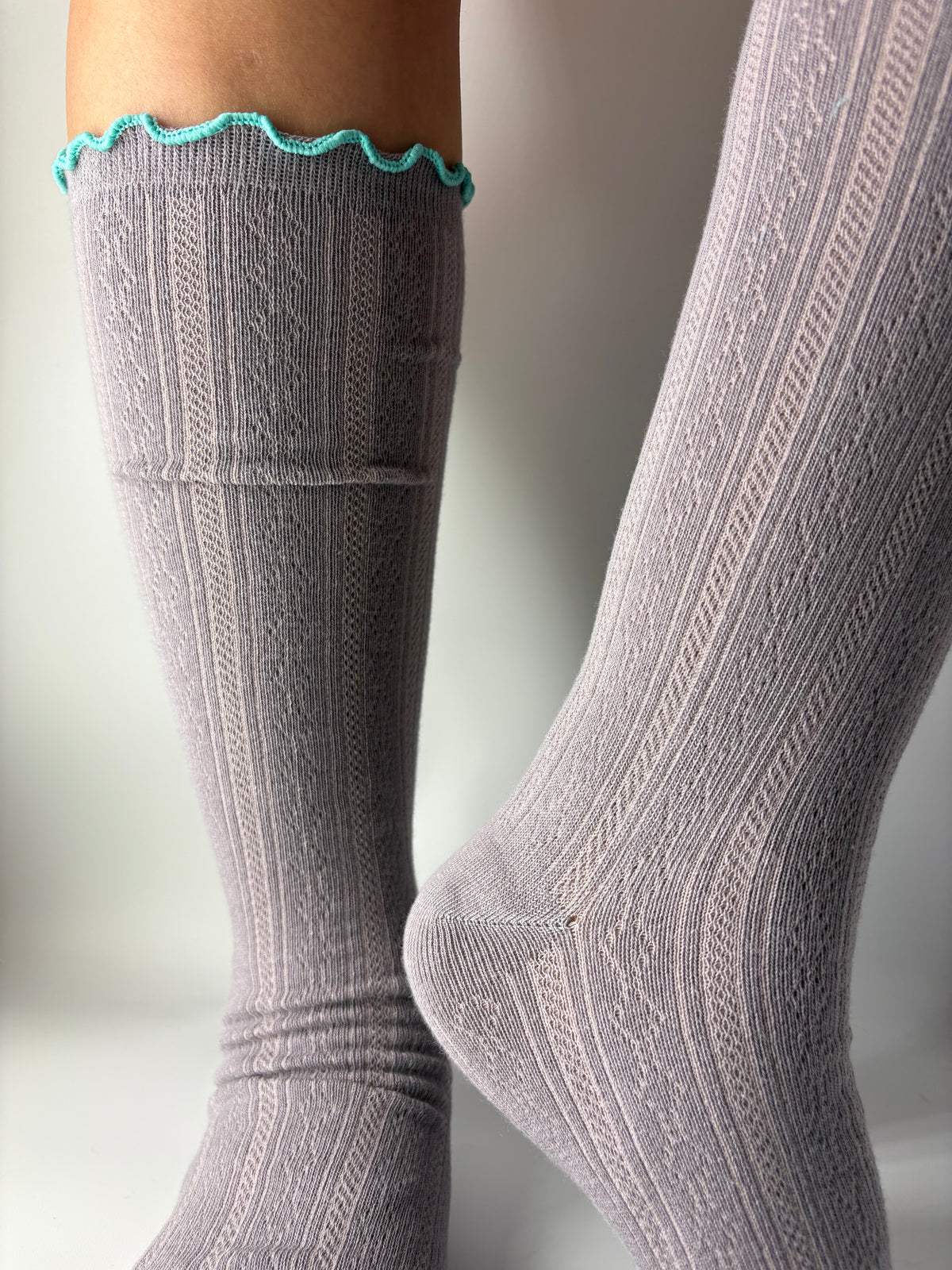 A pair of light gray pointelle knit knee-high socks with a bright turquoise blue scalloped trim at the top, shown worn on a person's legs against a neutral light gray background.