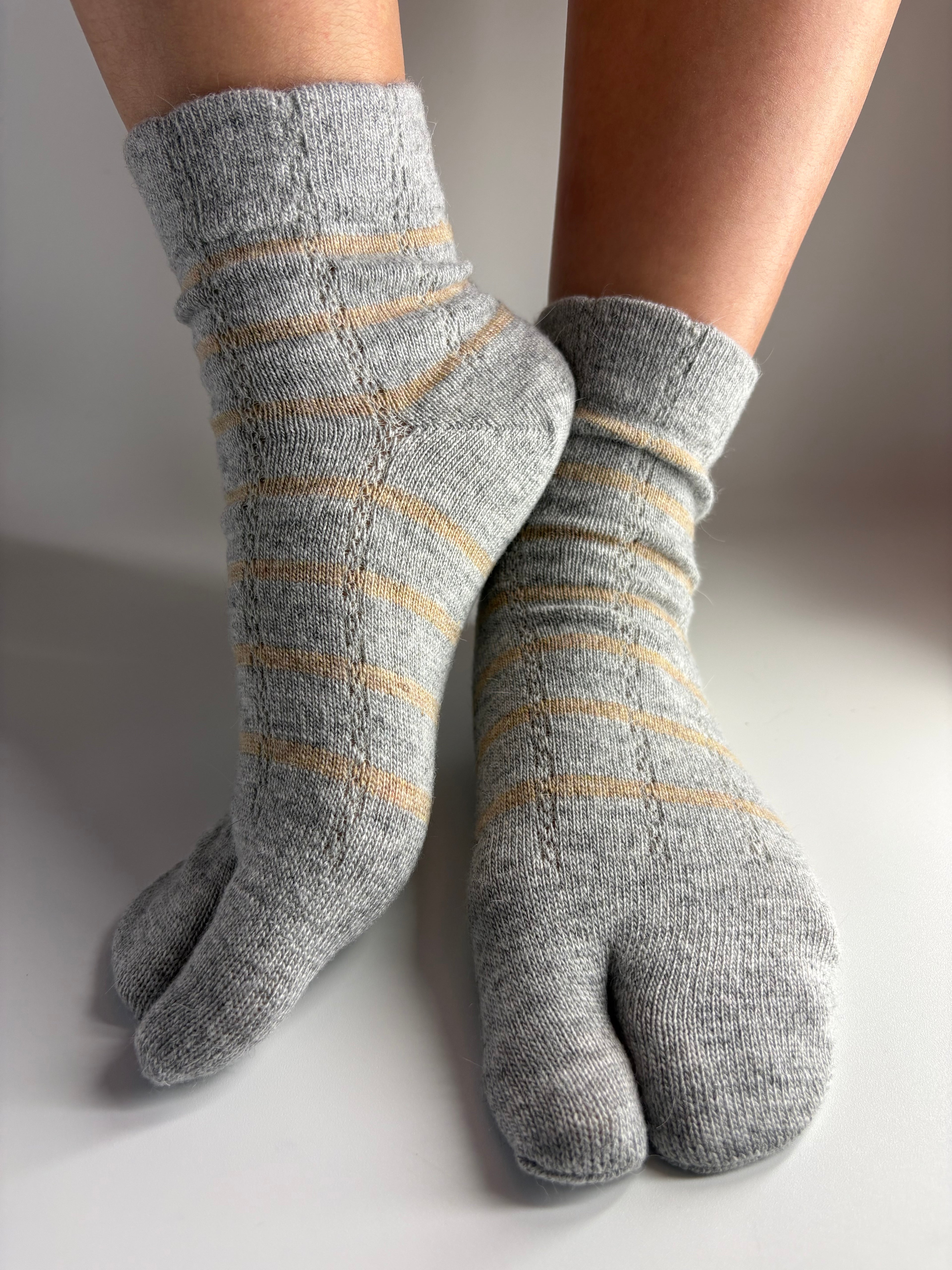 A pair of gray knit toe socks with thin horizontal tan stripes, shown worn on feet against a neutral light gray background.