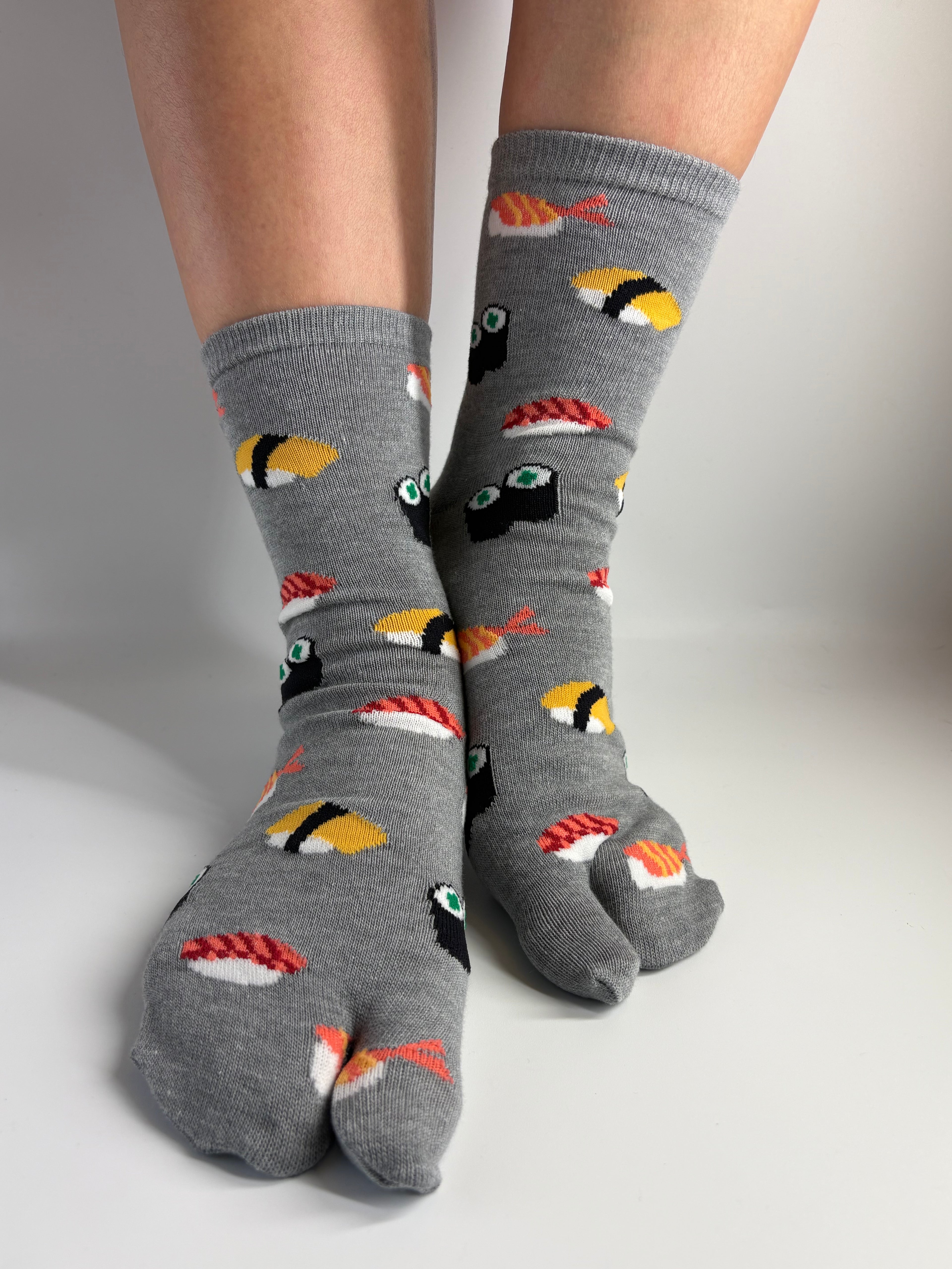 Gray tabi socks with a colorful sushi print, shown on a person's feet against a neutral studio background.