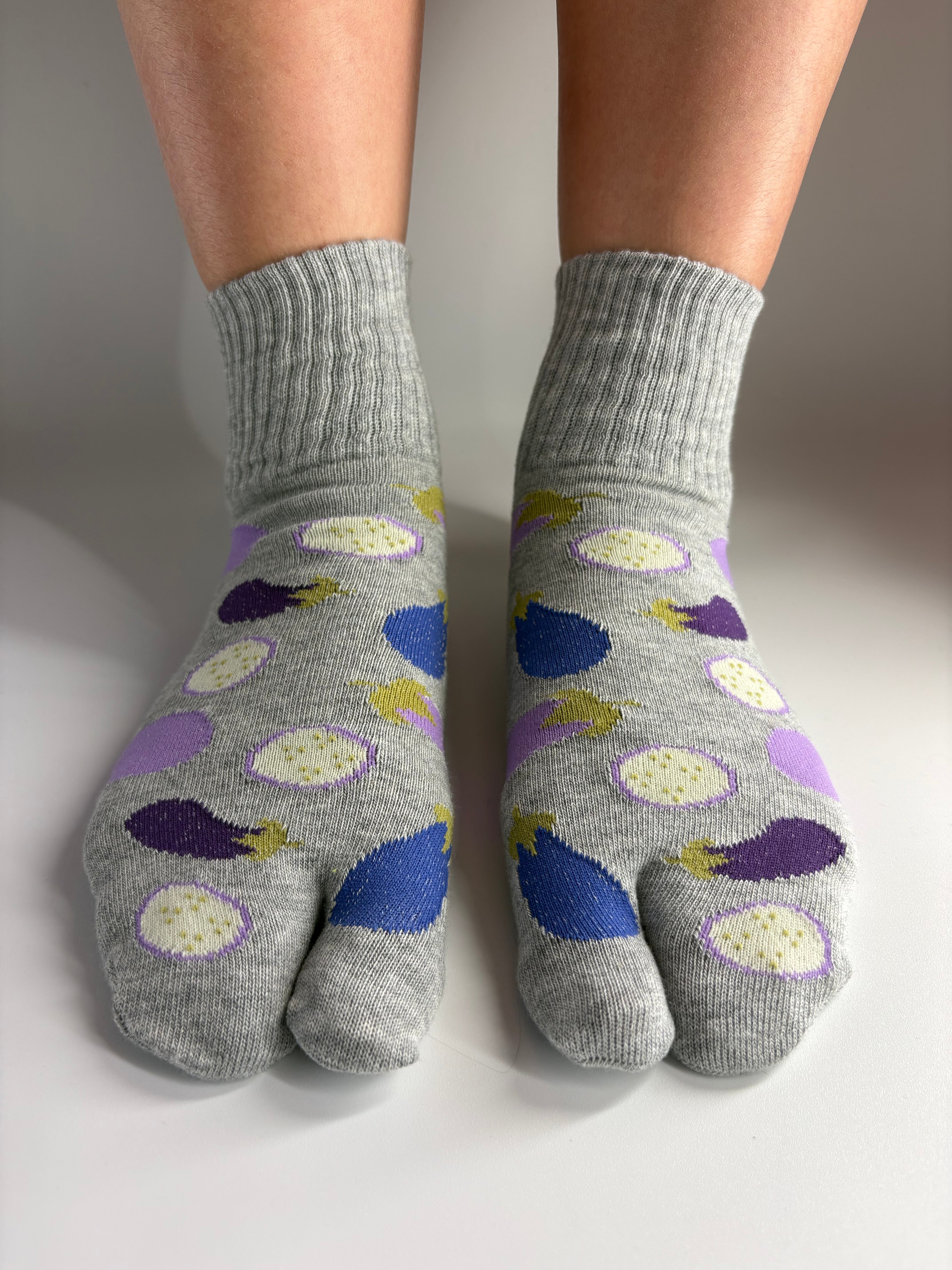 A pair of grey tabi socks with a pattern of purple eggplants,  shown on feet against a plain light background.