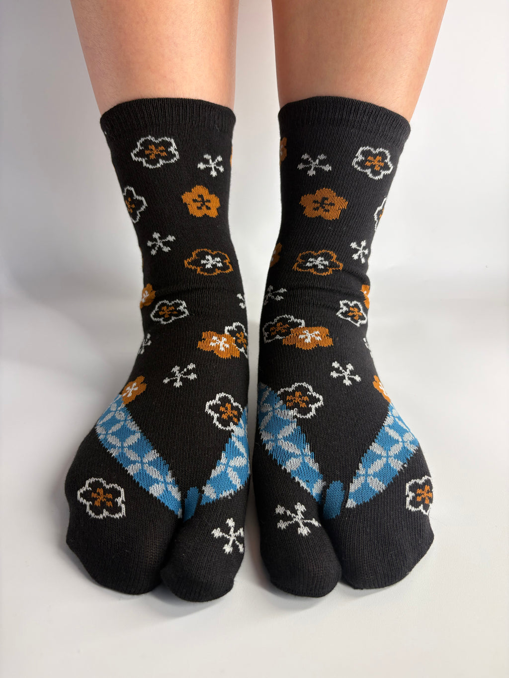 A pair of black  tabi socks with a pattern of white flowers, brown and gold blossoms, and light blue geometric accents, shown on feet against a plain white background.