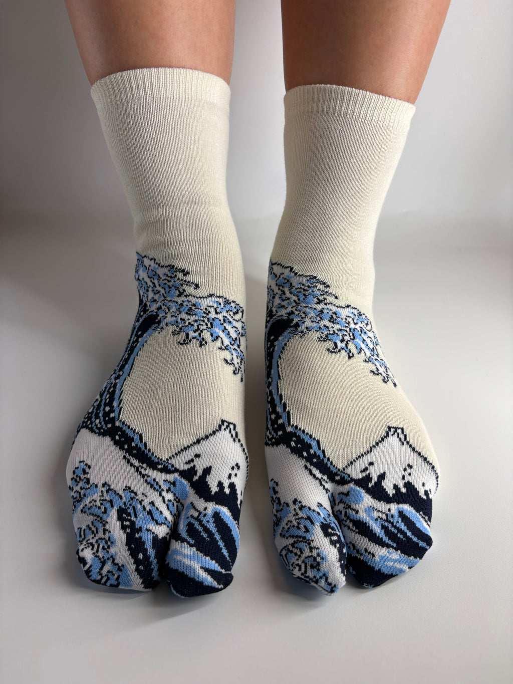 A pair of off-white crew socks with a blue and black great wave wave pattern, worn on feet against a plain light gray background.