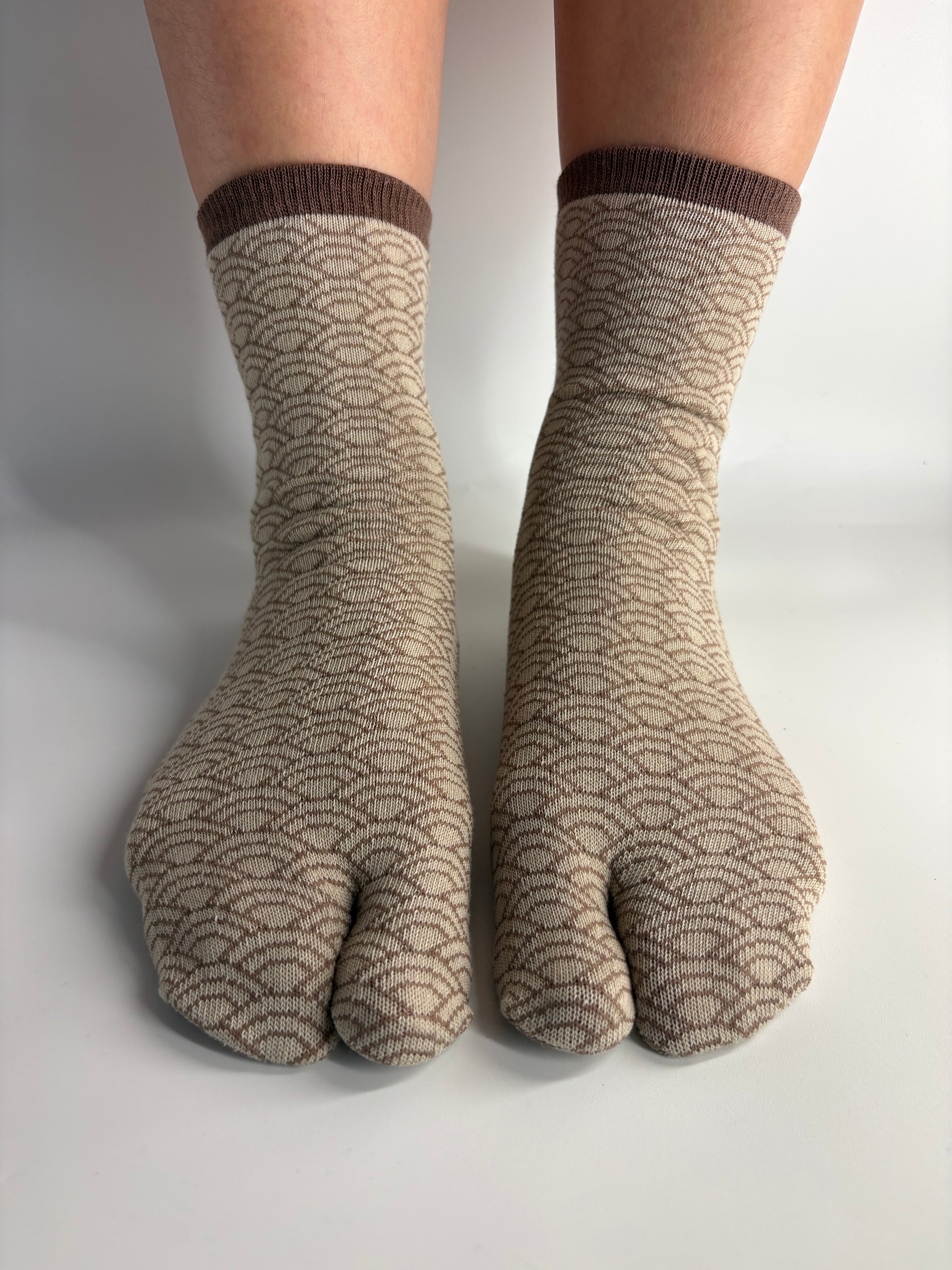 A pair of beige and brown seigeiha patterned tabi socks worn on feet, shown against a neutral light gray background.