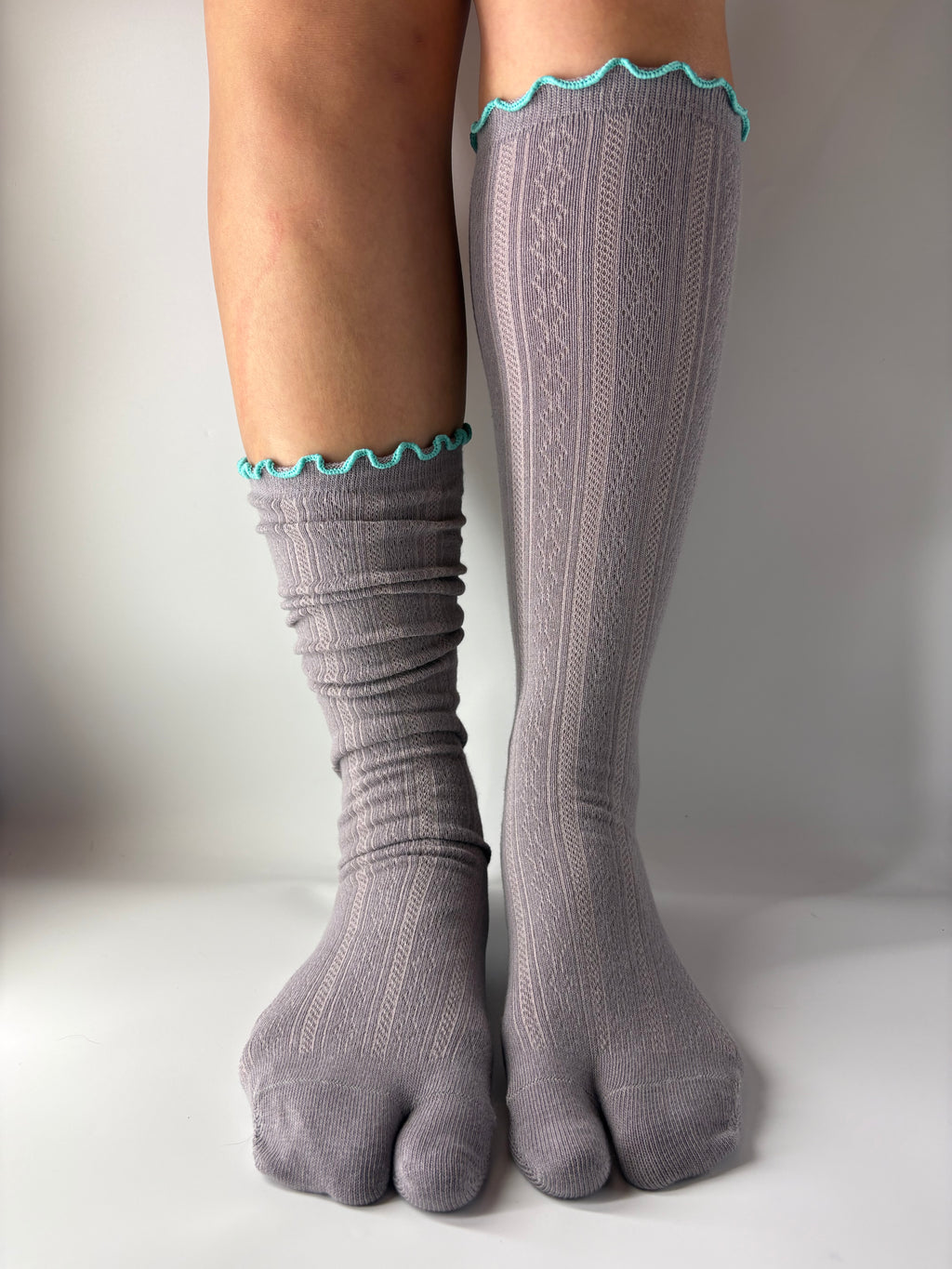 A pair of gray  pointelle open knit ribbed toe socks with a turquoise scalloped trim at the top, shown on a person's feet against a neutral gray background.