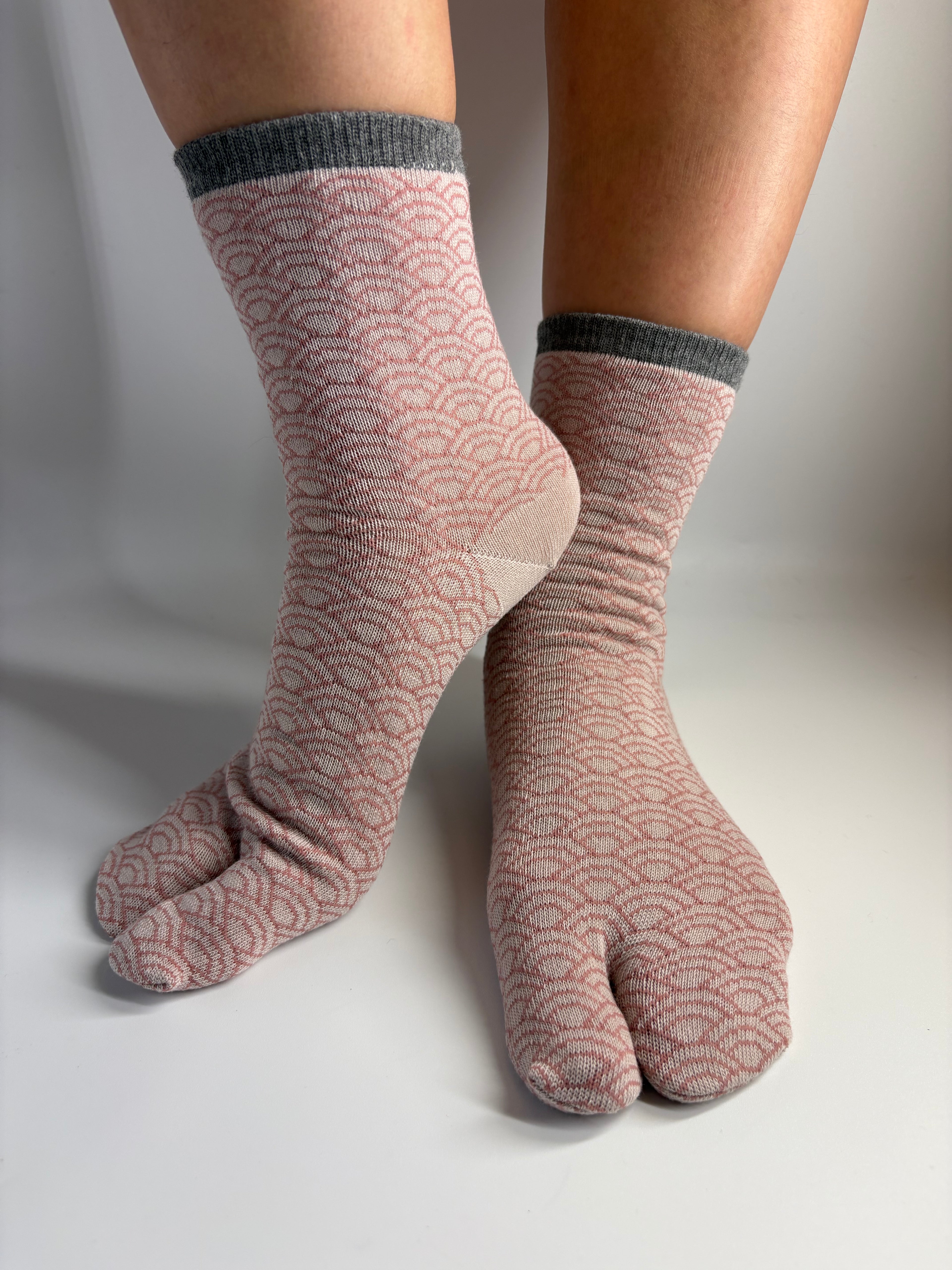 A pair of split-toe crew socks with a dusty pink and white wave pattern and dark gray ribbed cuffs, shown on a person's feet against a neutral studio background.