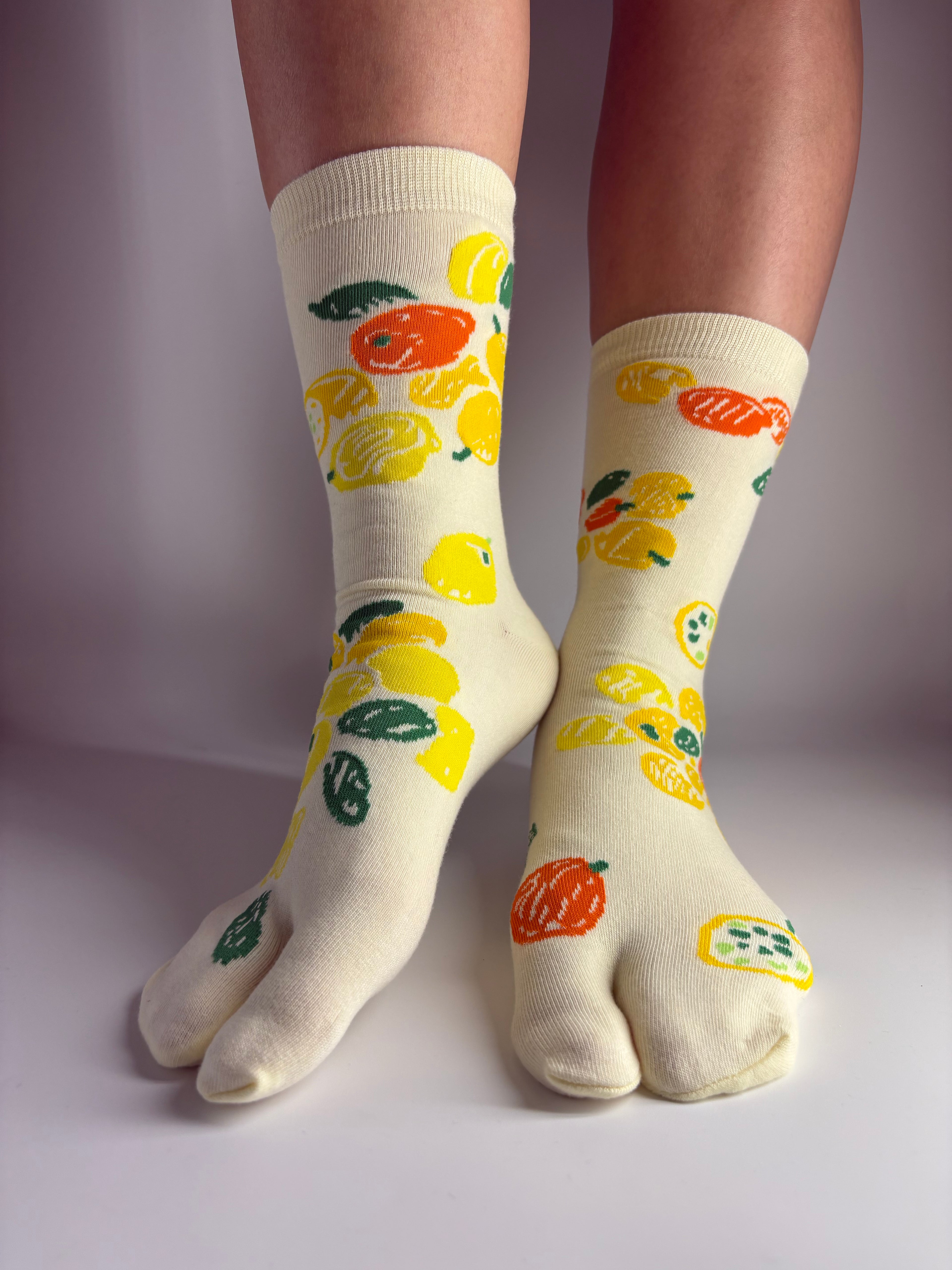 A pair of light yellow-colored crew socks with a colorful lemon and orange pattern, worn on feet against a plain light background.
