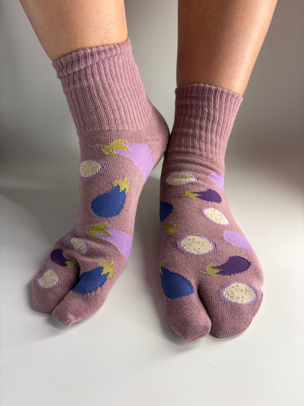 A pair of mauve-colored tabi socks with a pattern of purple eggplants a worn on feet against a neutral studio background.