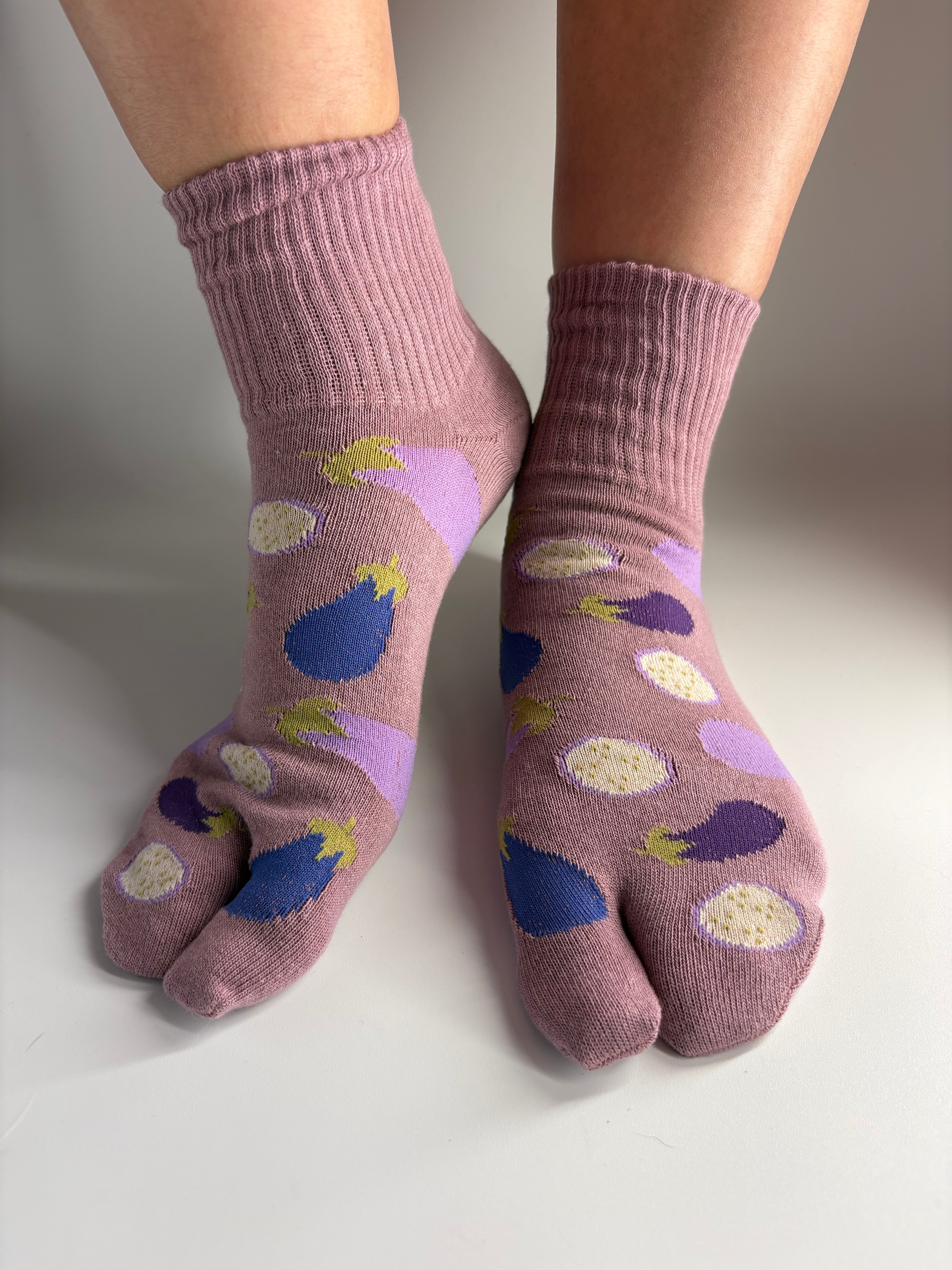 A pair of mauve-colored tabi socks with a pattern of purple eggplants a worn on feet against a neutral studio background.