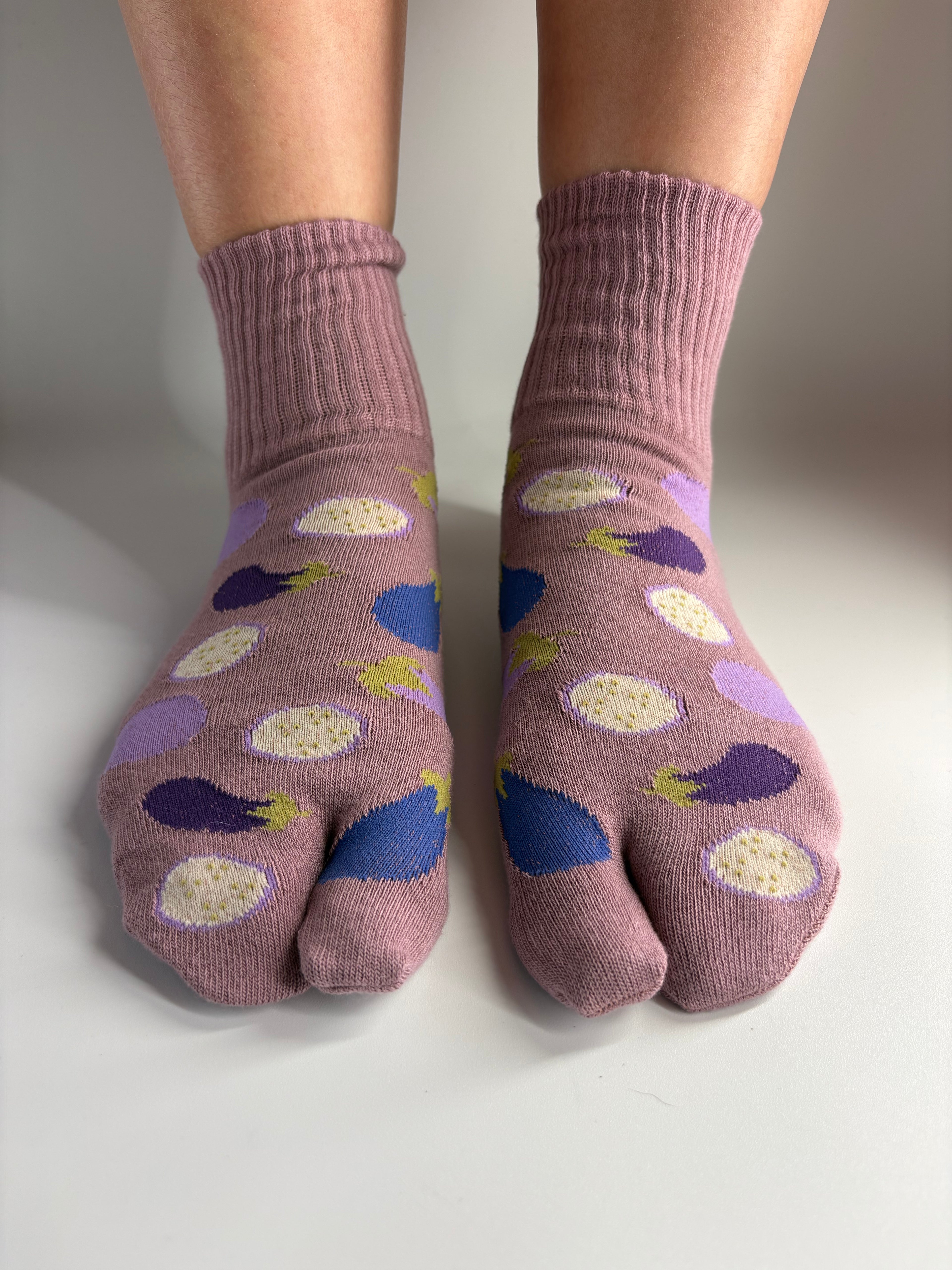 A pair of mauve-colored tabi socks with a pattern of purple eggplants, worn on feet against a neutral light gray background.