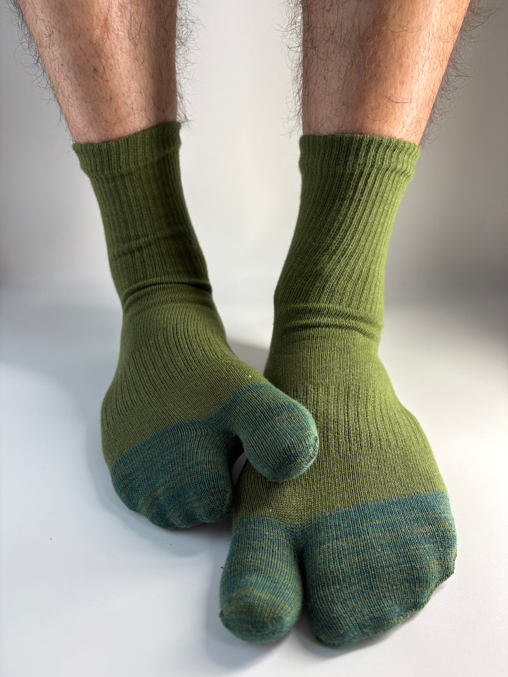 A pair of olive green and forest green split-toe socks worn on feet, shown against a plain light gray studio background.
