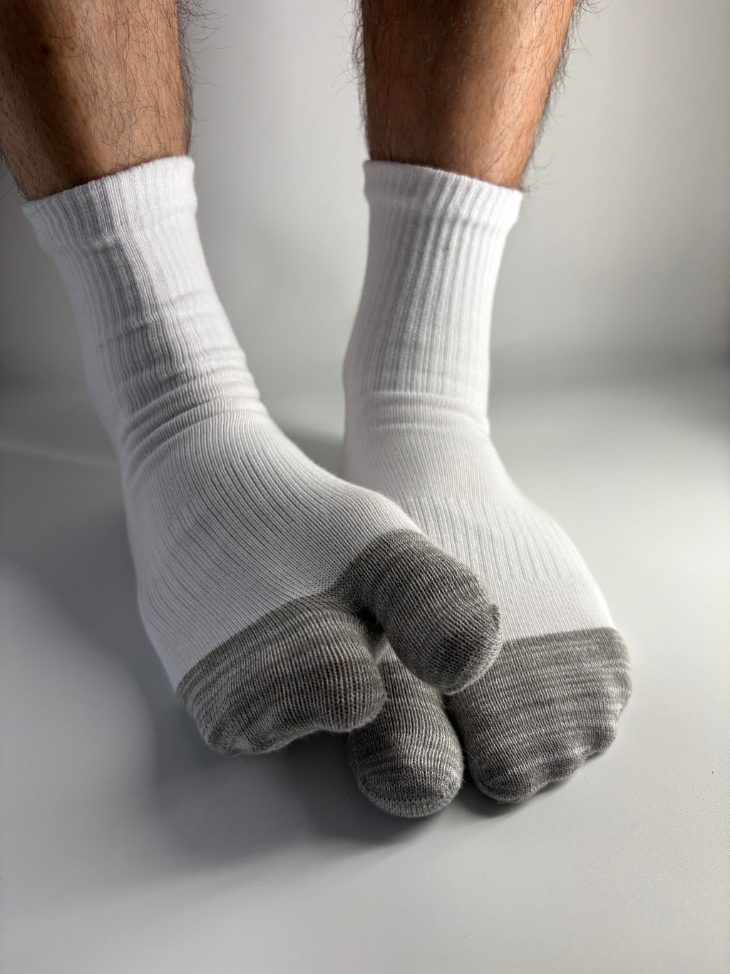 A close-up of a person's feet wearing white ribbed socks with gray toe sections, pressing down on a light gray surface.