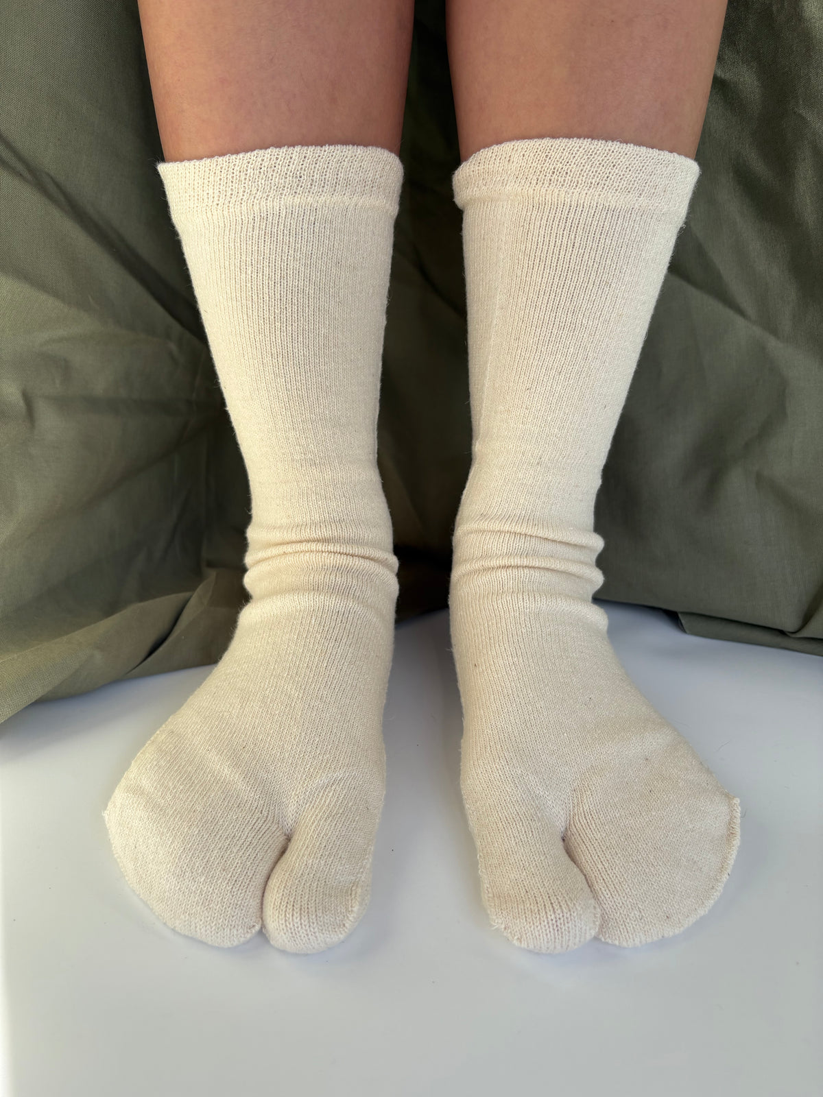 A pair of off-white Japanese tabi socks worn on feet, shown against a background of olive green fabric and a white surface.
