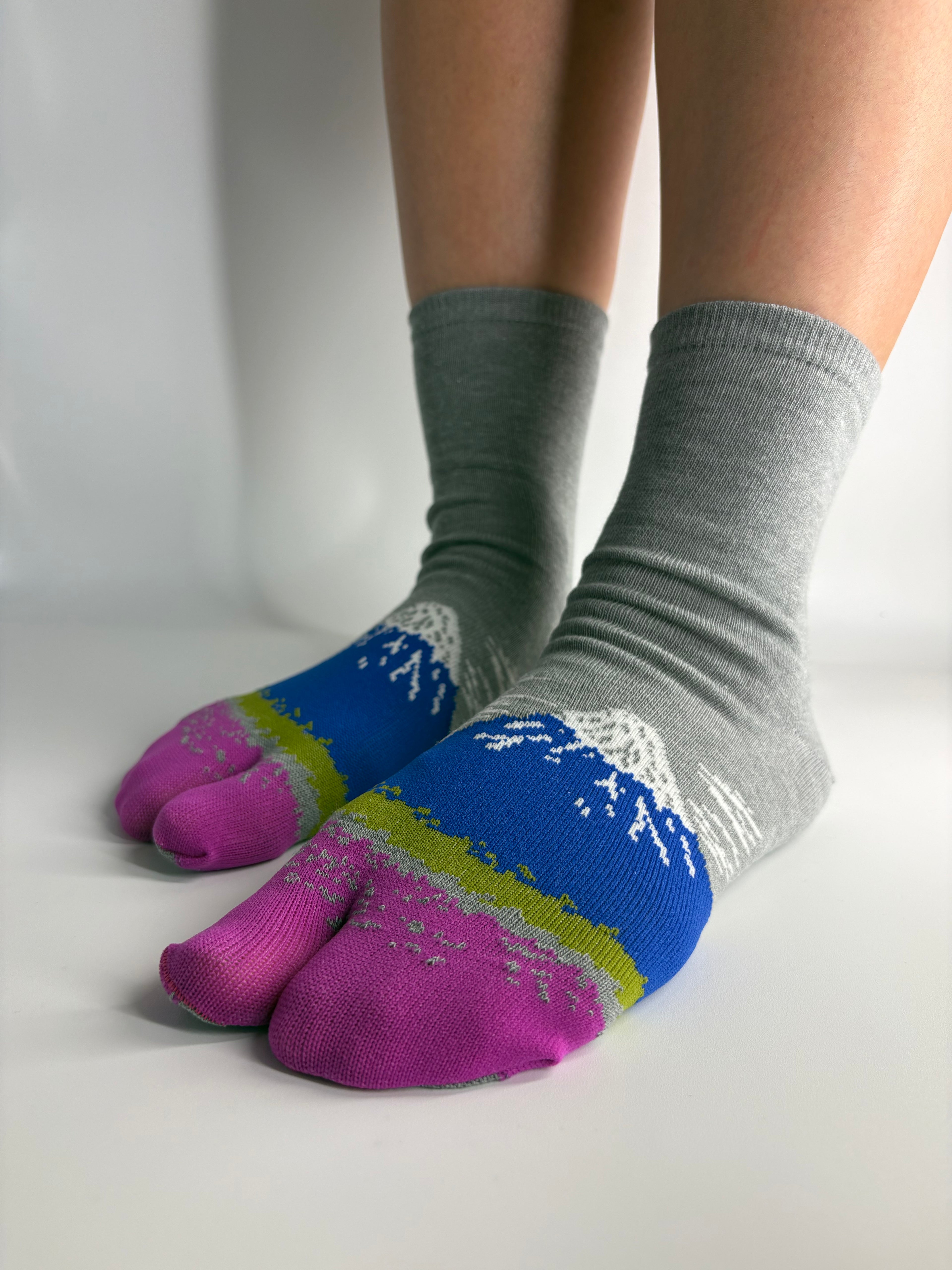 A pair of split-toe socks worn on feet resting against a white wall, showing a knit pattern with bright pink toes, blue and gray sections, and a lime green band all representing mount fuji in japan