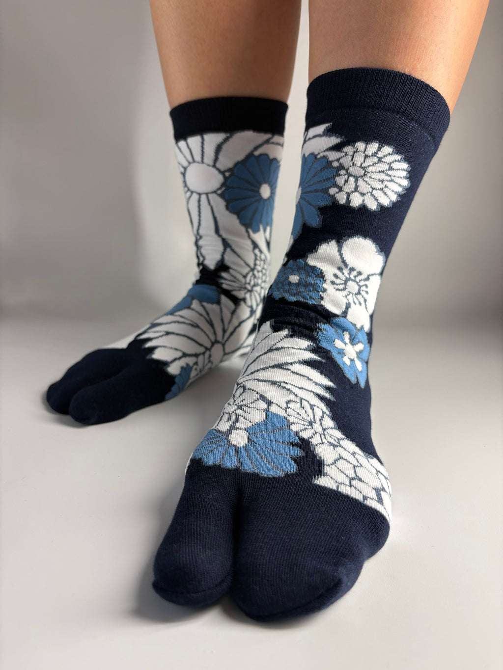 A pair of split-toe socks worn on feet, featuring a navy blue base with a white and light blue floral pattern, set against a plain light gray background.