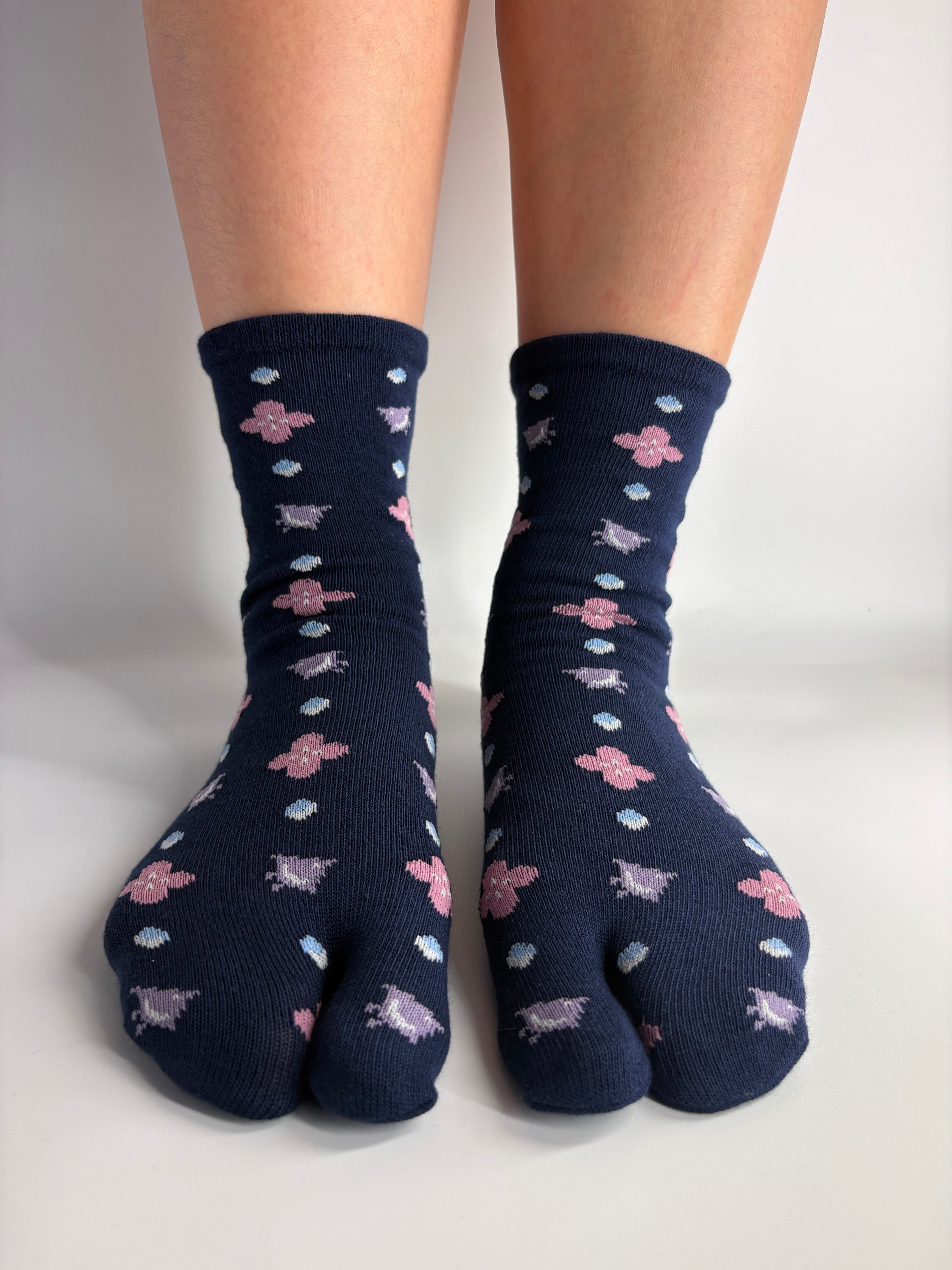 A pair of dark navy split-toe socks with a repeating pattern of pink flowers, lilac birds, and small light blue and white dots, shown on feet against a plain white background.
