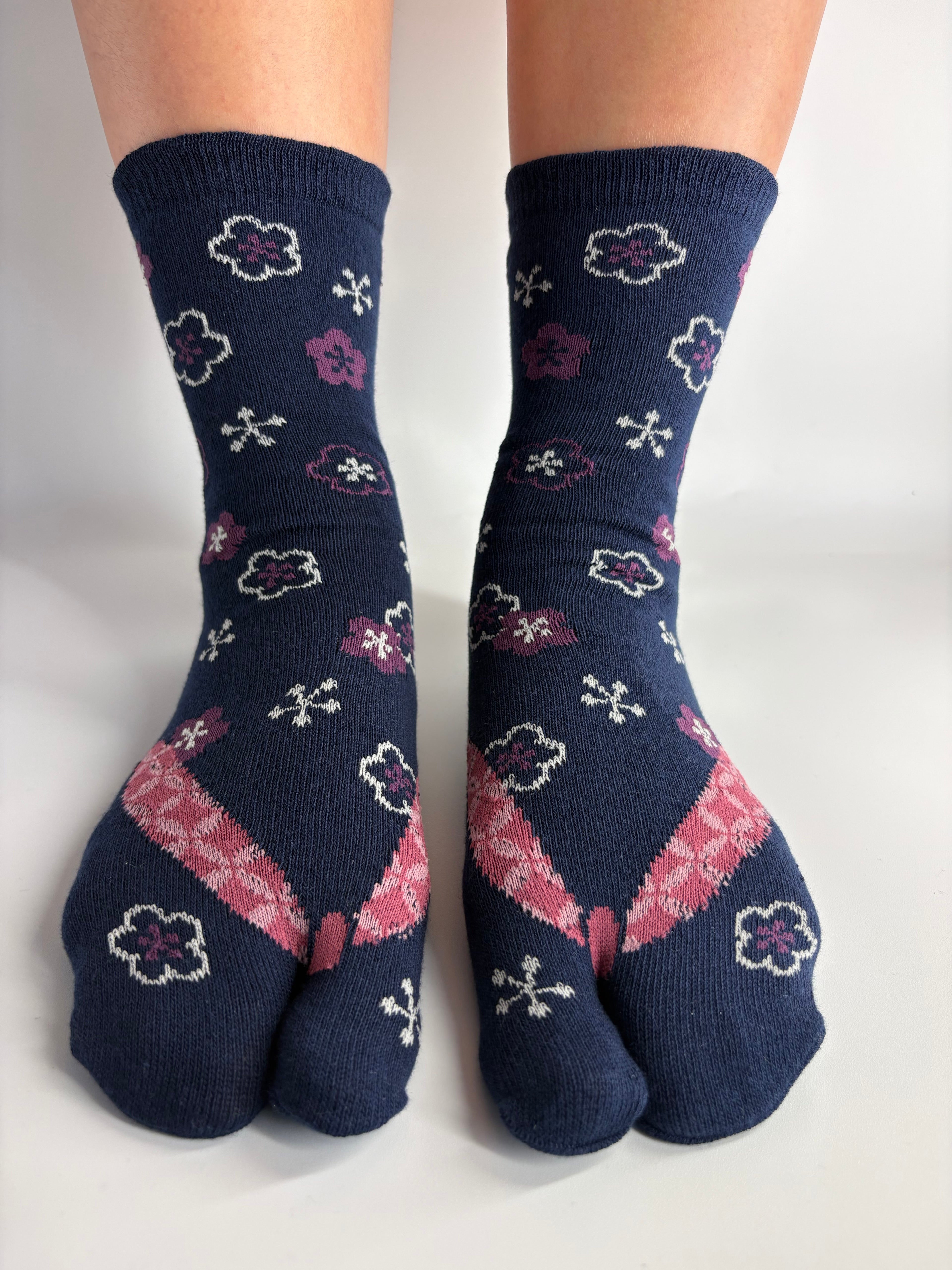A pair of navy blue split-toe socks with a pattern of white and purple flowers, pink and purple geometric shapes, and small white cross motifs, in the print of geta shown worn on feet against a plain white background.