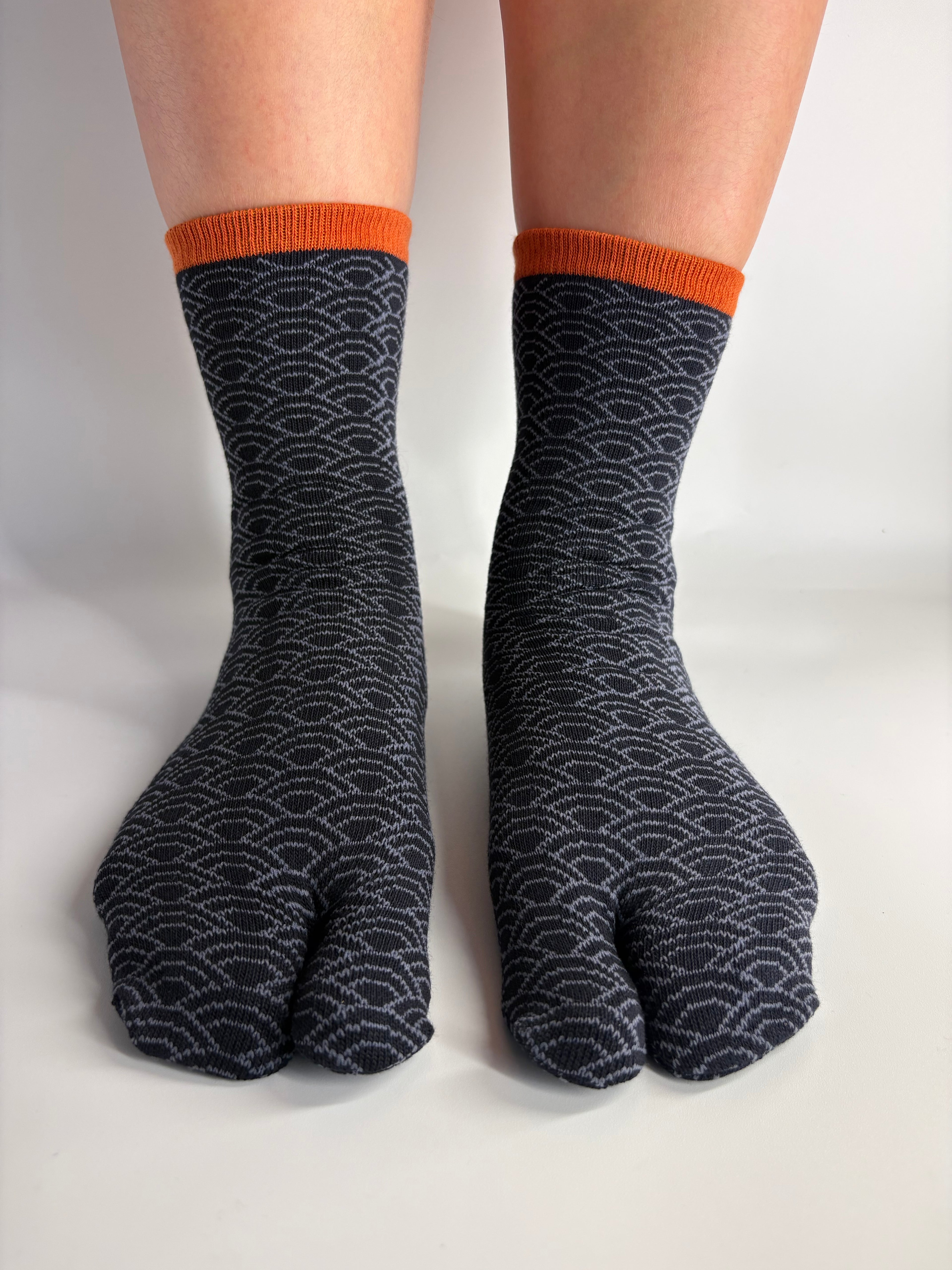 A pair of dark navy tabi socks with a traditional japanese pattern and orange ribbed cuffs, shown on feet against a plain white background.