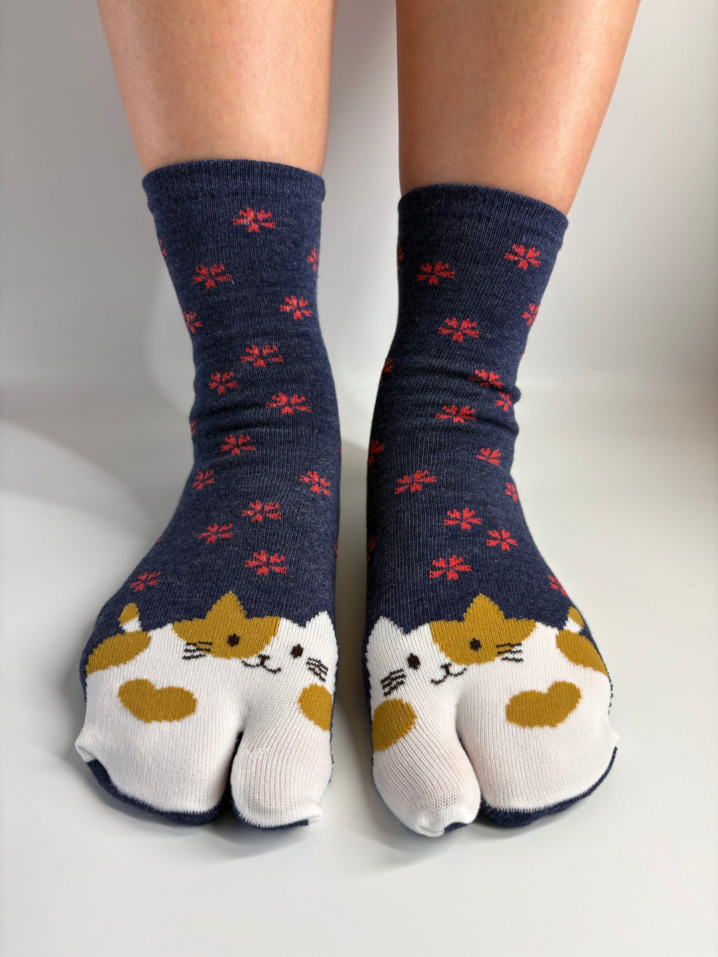 A pair of navy blue tabi socks with a pattern of small red flowers and cat faces on the toes, worn on a person's feet against a neutral light gray background.