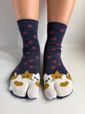 A pair of navy blue tabi socks with a pattern of small red flowers and cat faces on the toes, worn on a person's feet against a neutral light gray background.
