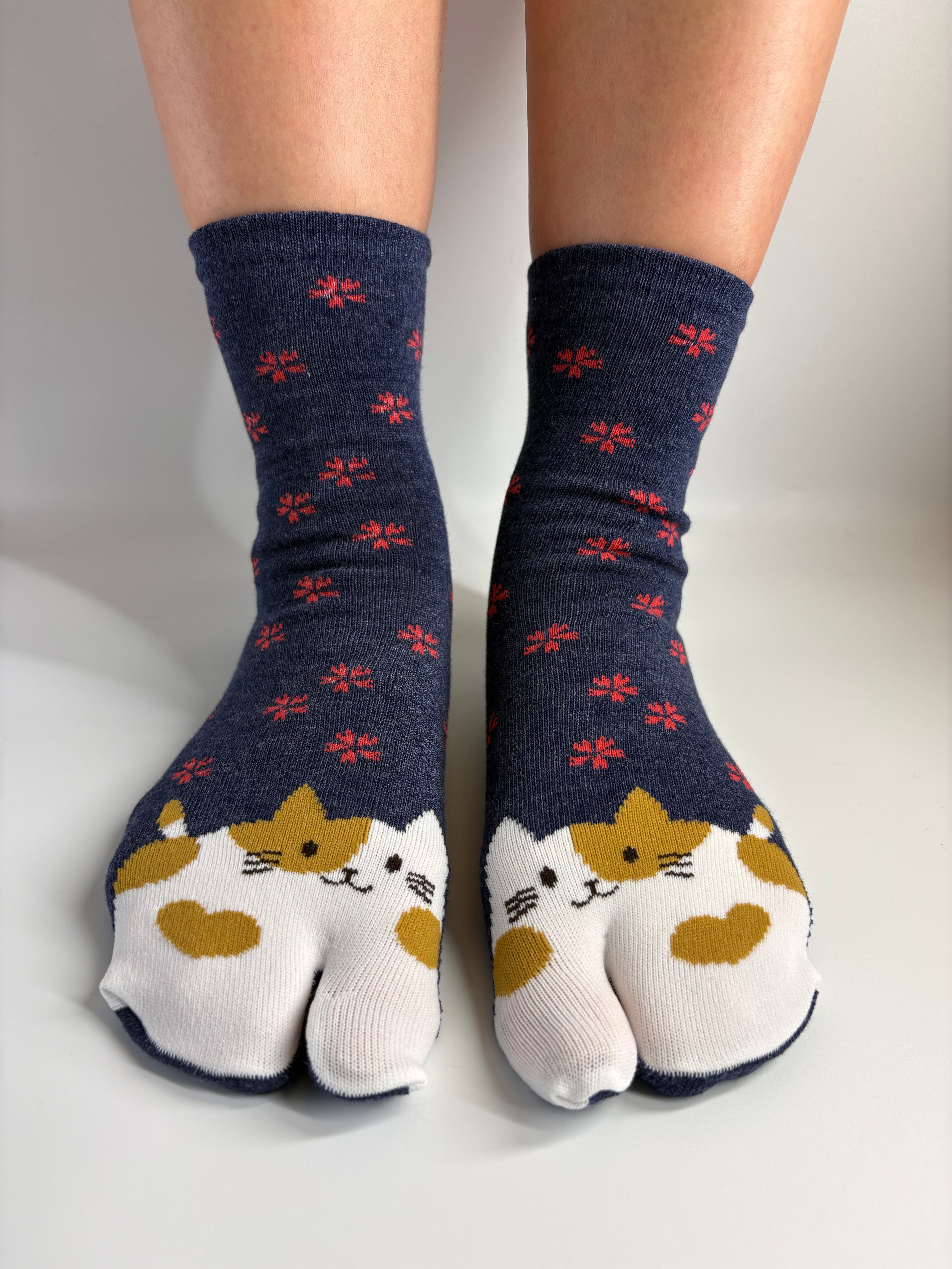 A pair of navy blue tabi socks with a pattern of small red flowers and cat faces on the toes, worn on a person's feet against a neutral light gray background.