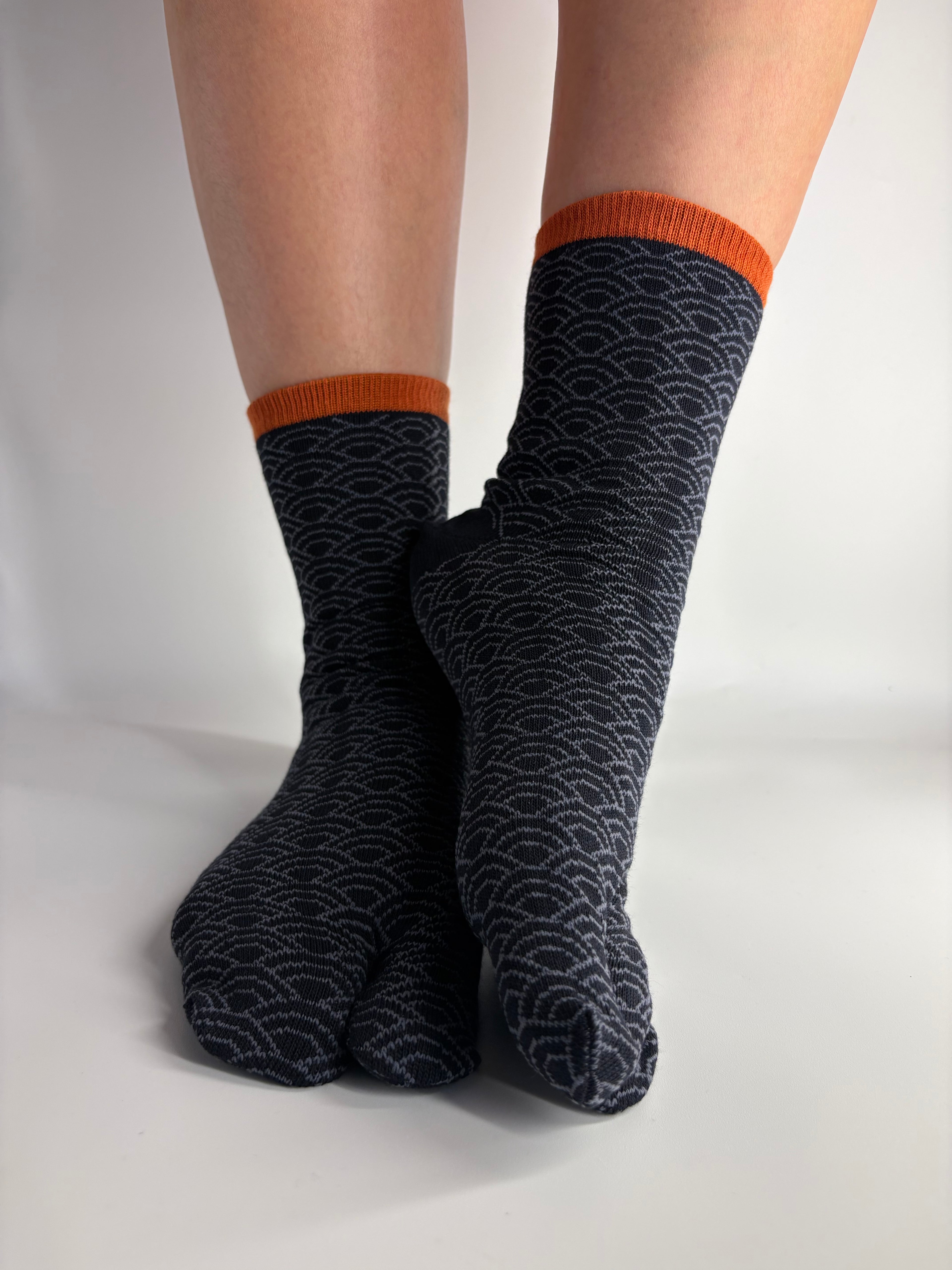 A pair of dark navy segeiha print crew socks with a light gray wave pattern and orange ribbed cuffs, worn on feet resting against a white background.