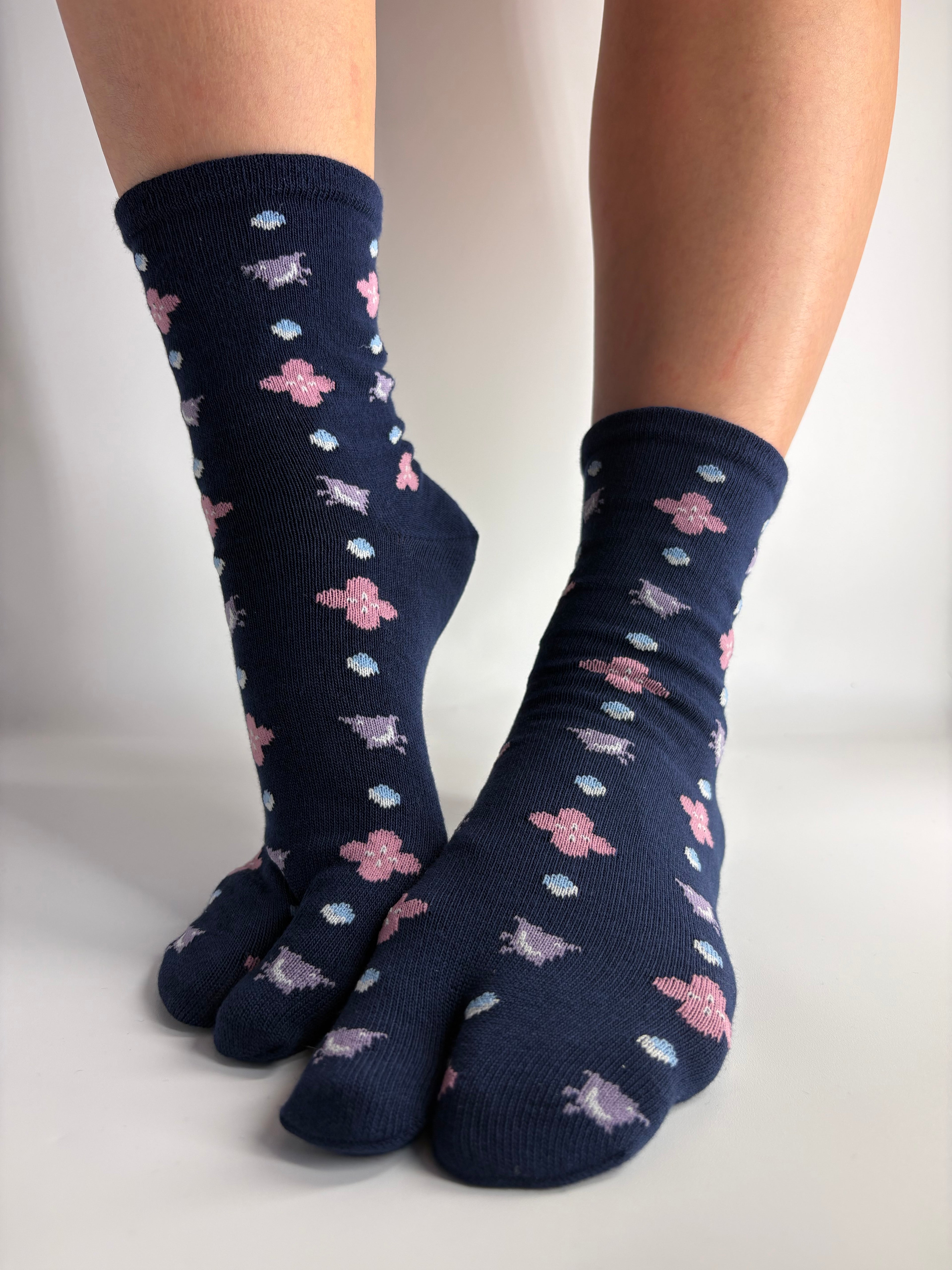 A pair of dark navy split-toe socks with a repeating pattern of pink flowers, lilac birds, and small light blue and white dots, shown on feet against a plain white background.