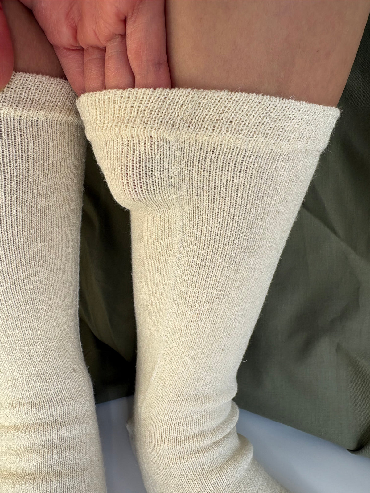Close-up of a person wearing off white tabi knit socks, with one leg pulled up to show the cuff texture. A hand is visible at the top, and an olive green garment is in the background.