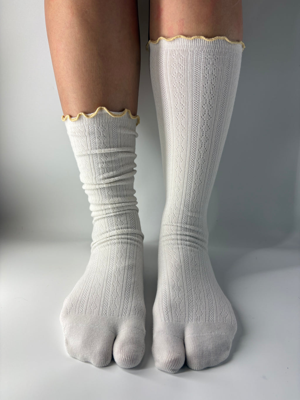 A pair of off-white ribbed tabi split toe socks with a lettuce edge yellow trim, shown on a person's legs against a neutral gray background.