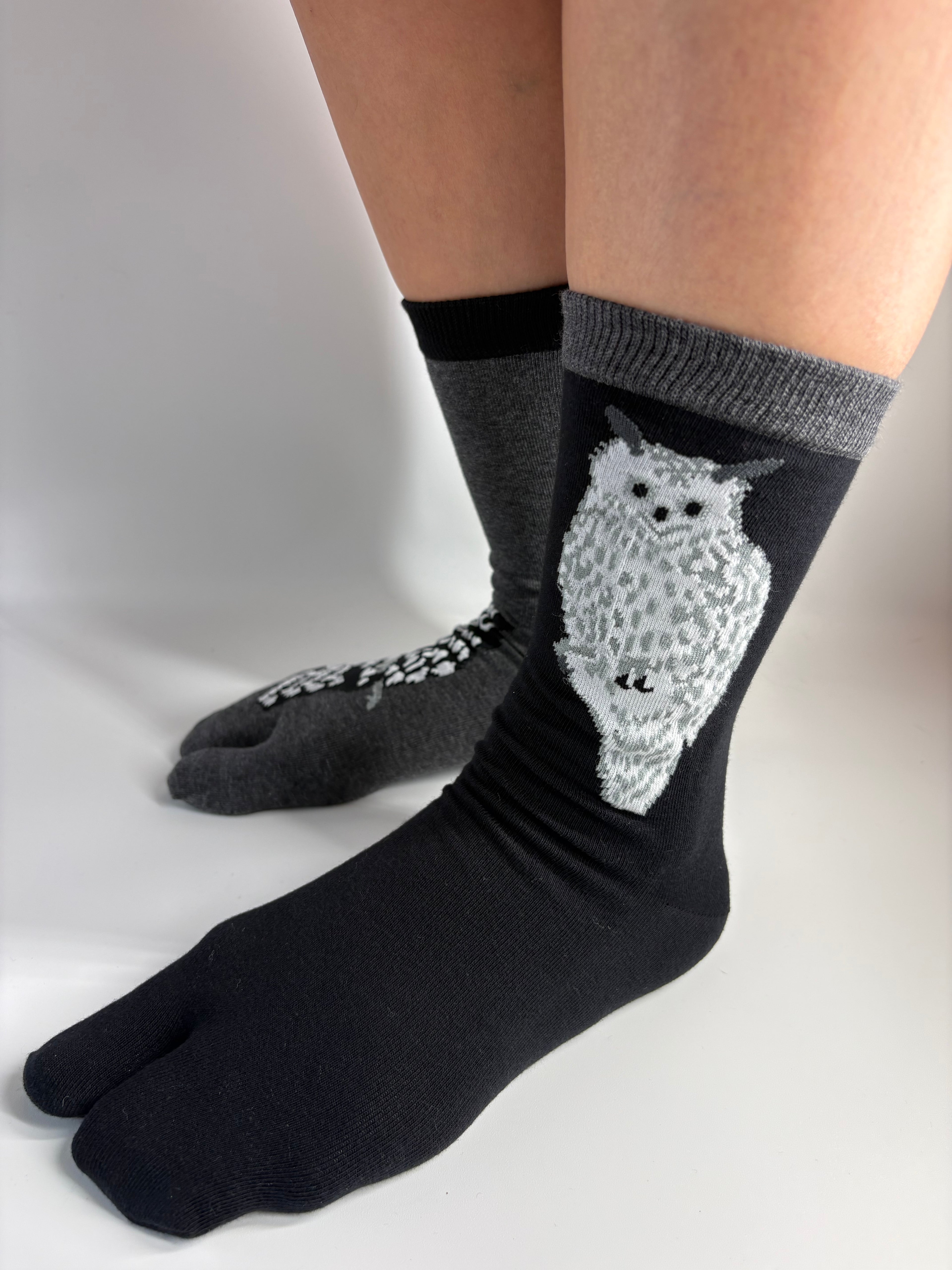 A pair of black and gray socks worn on feet, featuring a white and gray spotted owl graphic on the front sock.