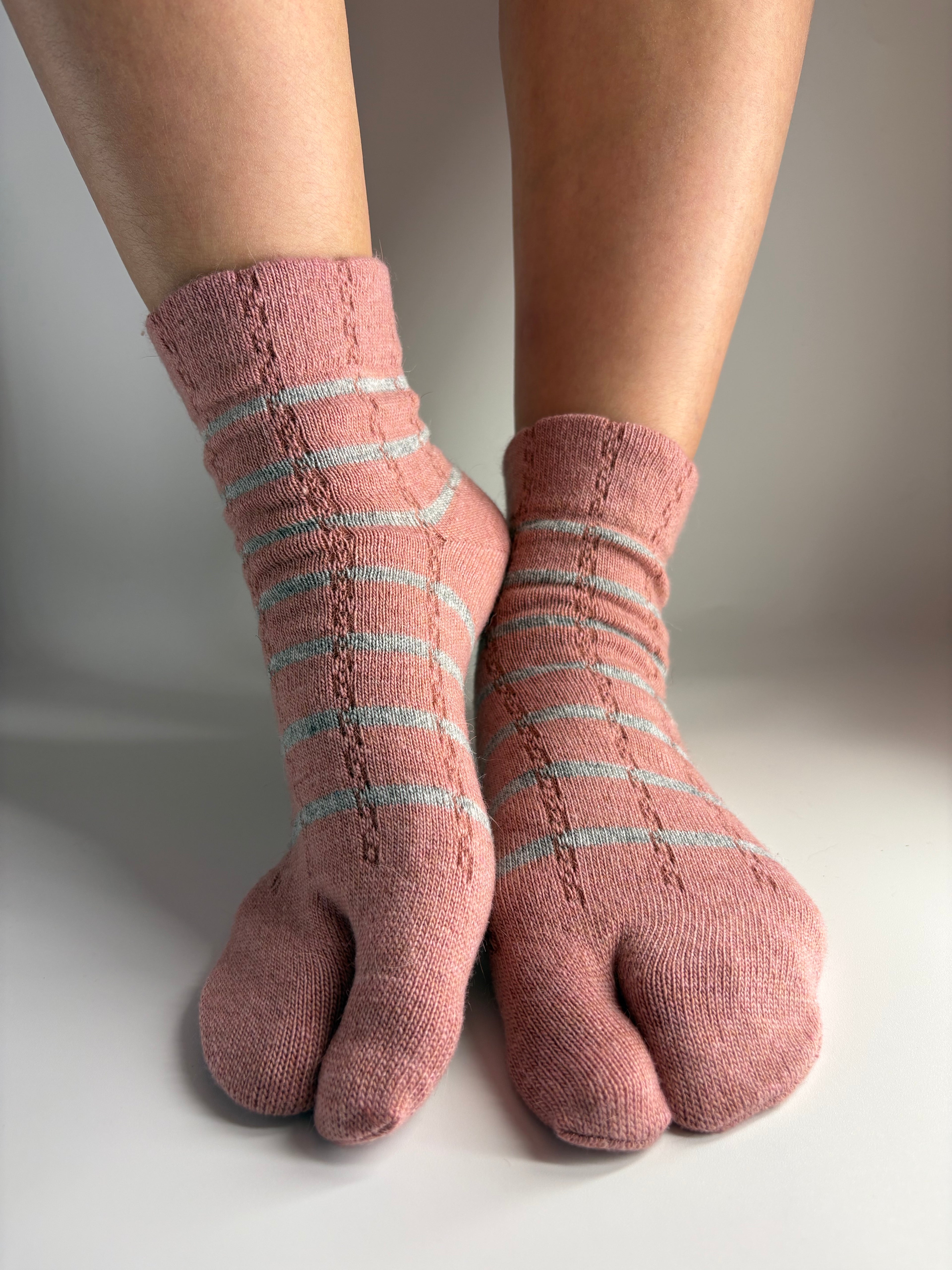 A pair of pink tabi socks with light grey stripes, worn on a person's feet against a plain light gray background.