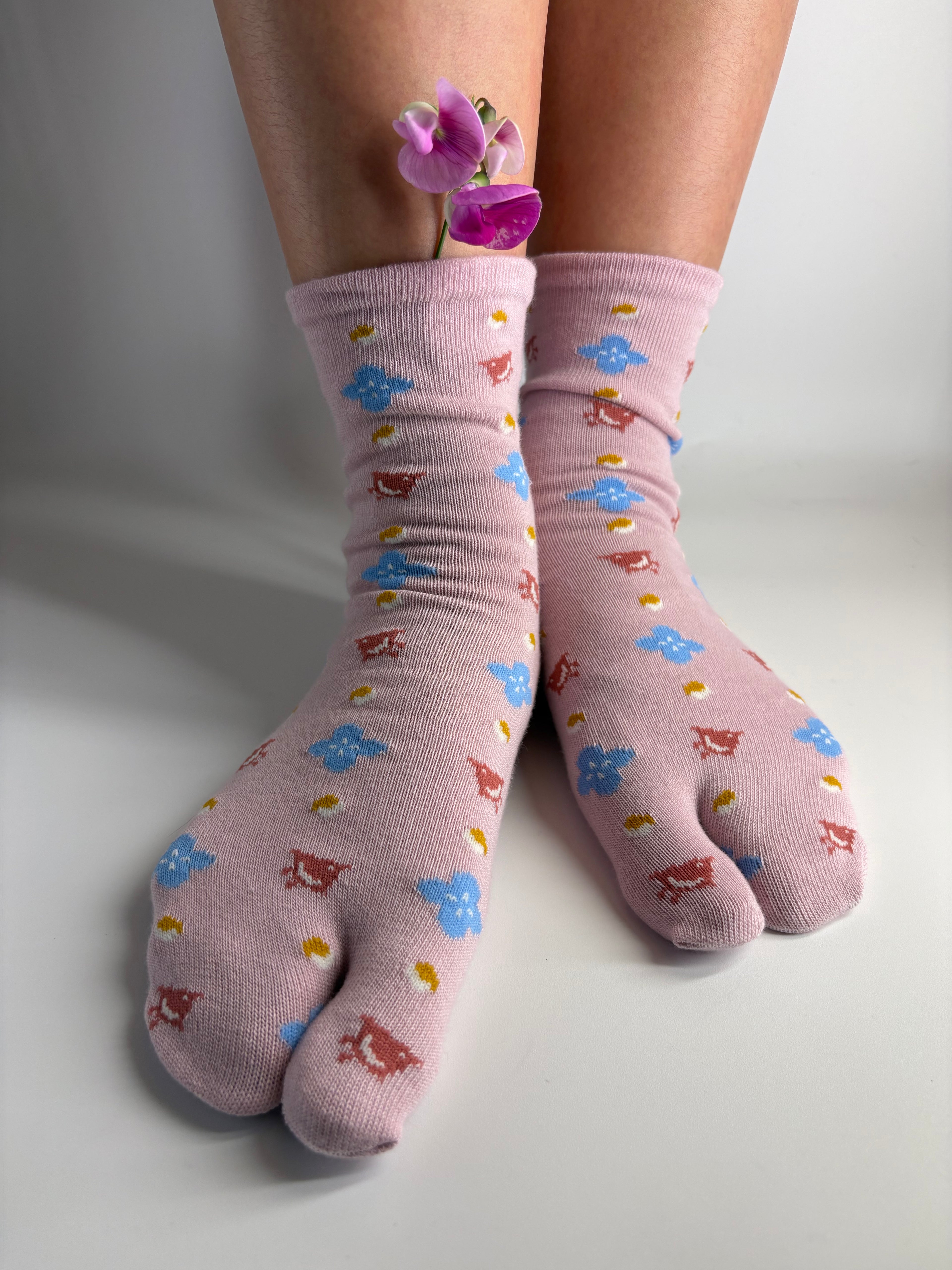 A pair of light pink tabi socks with a pattern of small blue, brown, and yellow japanese motifs (florals and birds) worn on feet with a purple orchid flower tucked into one ankle to style