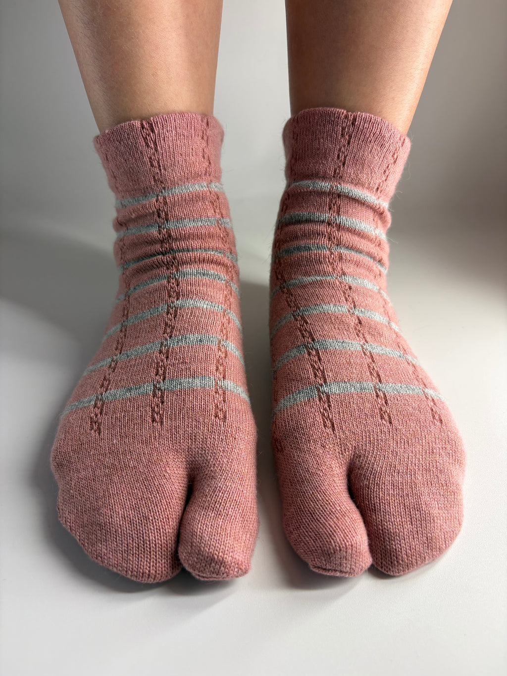 A pair of pink tabi socks with light grey horizontal stripes, shown on a person's feet against a neutral gray background.