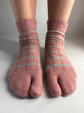 A pair of pink tabi socks with light grey horizontal stripes, shown on a person's feet against a neutral gray background.