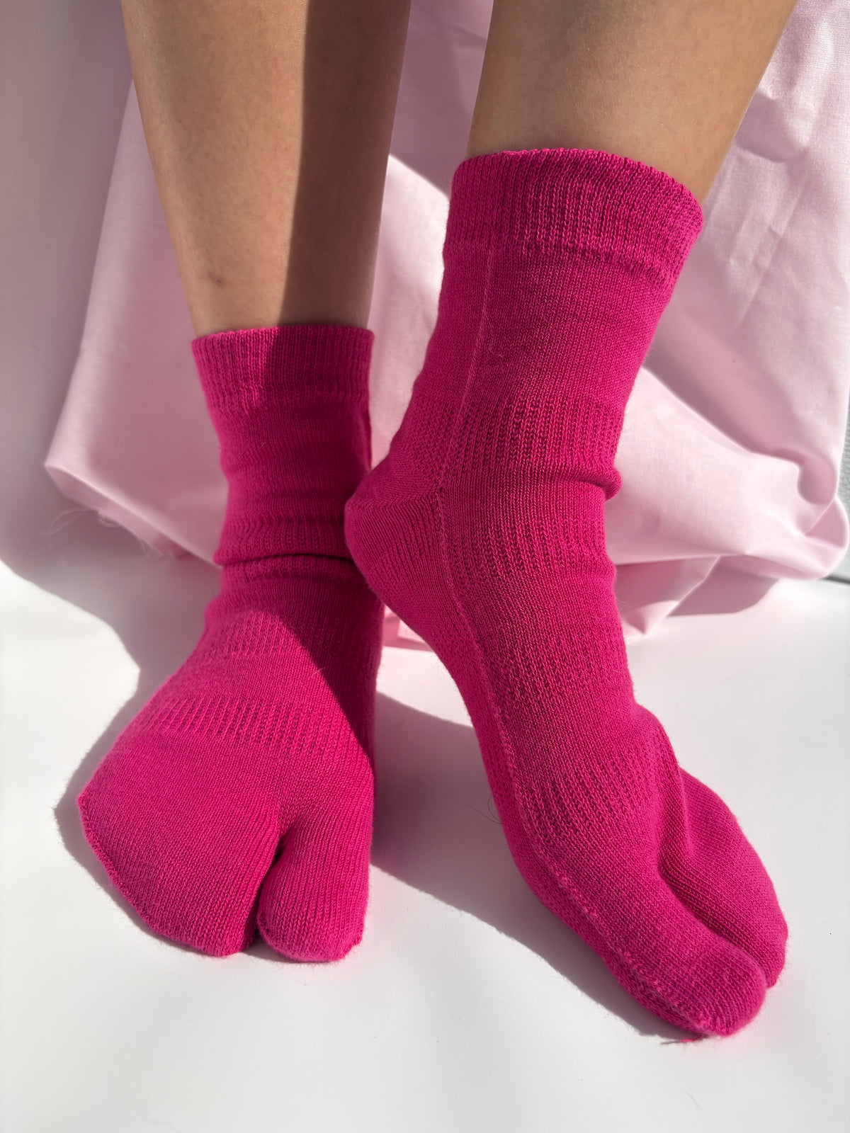 fuchsia tabi socks worn on a person's feet, shown against a soft light pink fabric backdrop on a white surface.