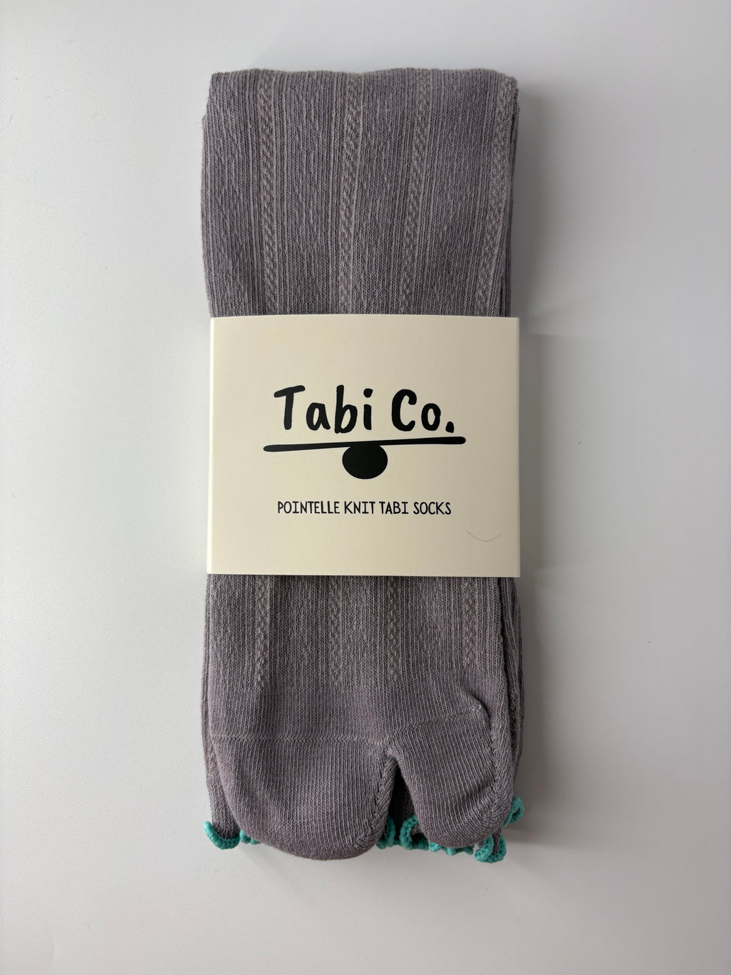 A pair of folded gray knit tabi socks with a white paper band label packaging that reads 'Tabi Co. POINTELLE KNIT TABI SOCKS', photographed against a plain light gray background.