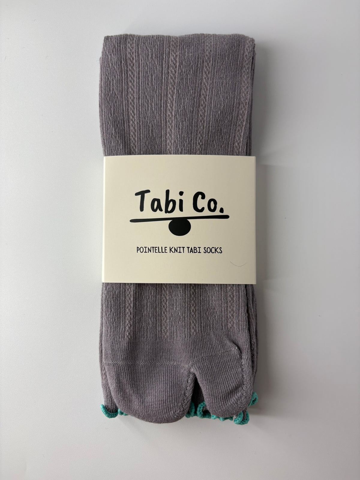 A pair of folded gray knit tabi socks with a white paper band label packaging that reads 'Tabi Co. POINTELLE KNIT TABI SOCKS', photographed against a plain light gray background.
