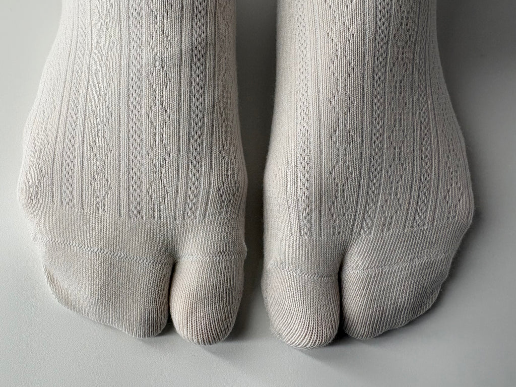 A close-up of a person's feet wearing off-white ribbed knit tabi socks, showing the textured knit pattern and toe separation on a neutral gray background.