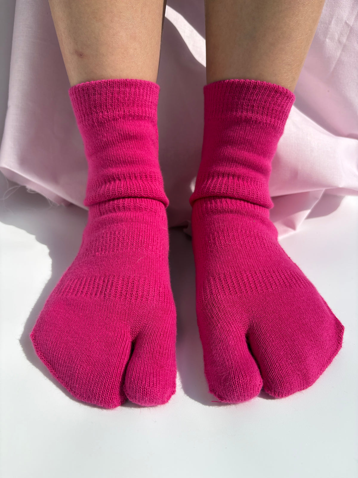 Bright pink tabi socks worn on a person's feet, shown against a white surface with light pink fabric in the background.