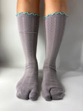 A pair of gray ribbed split toe socks with a turquoise scalloped trim at the top, pointelle knit shown on a person's feet against a plain light gray background.